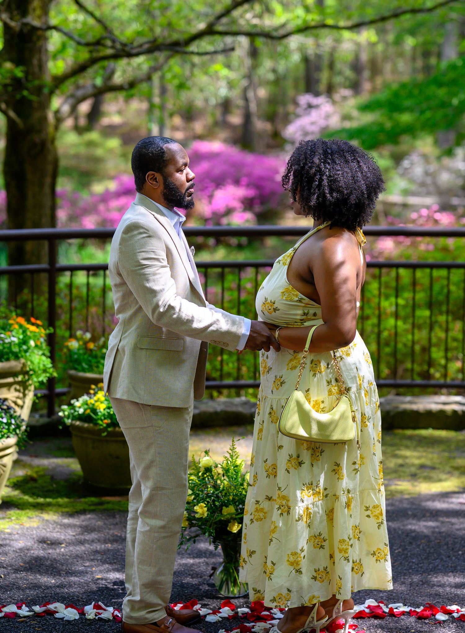 A couple holds hands tenderly in a lush garden with blooming pink azaleas, man in beige suit, woman in yellow floral dress.