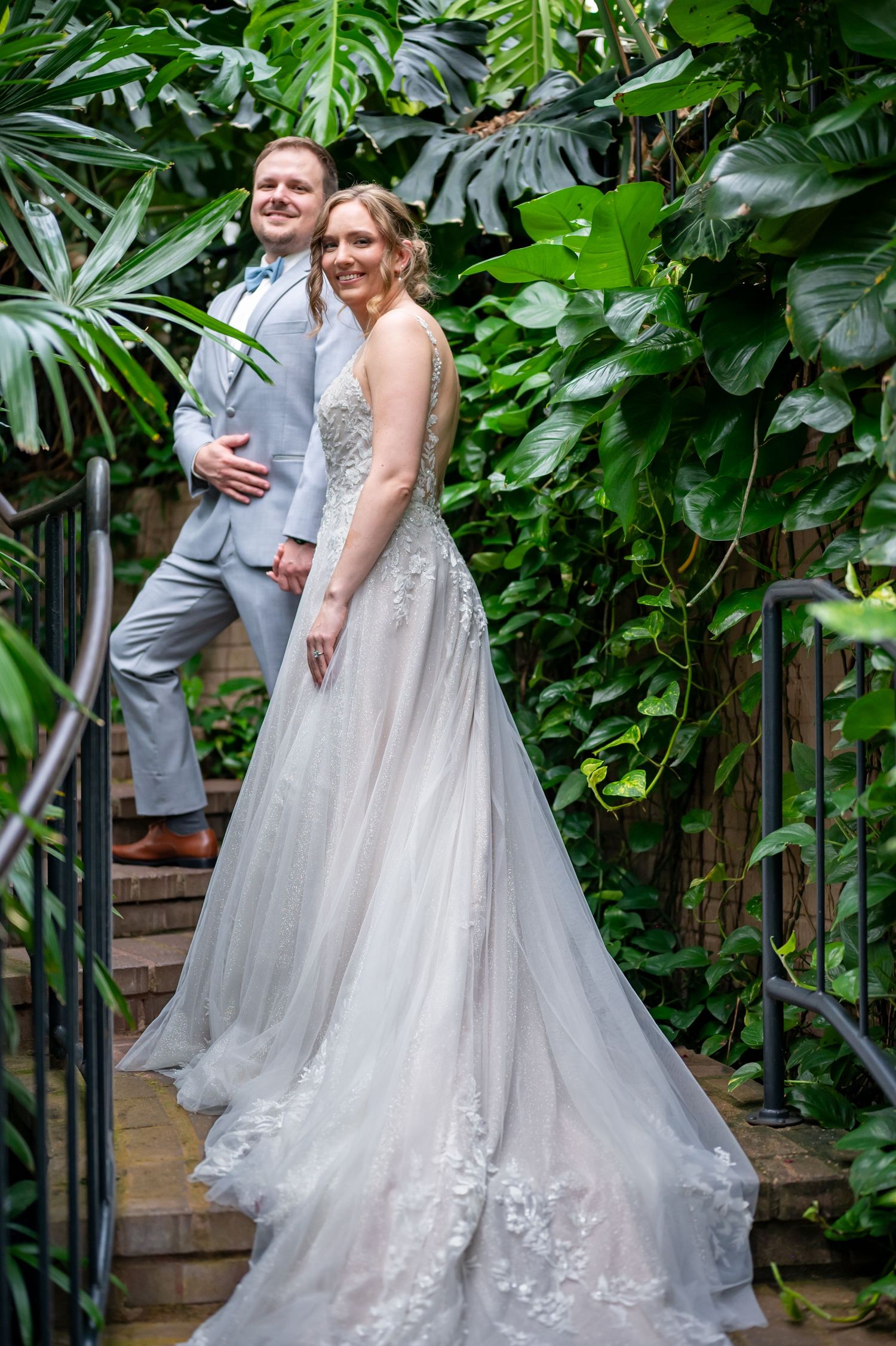 A couple in wedding attire pose together on brick steps surrounded by lush green tropical plants and foliage.