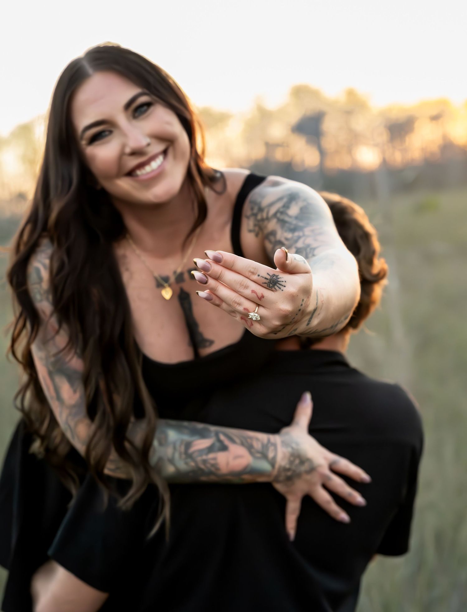 A woman in black shows off her tattoo sleeves while posing outdoors during golden hour.