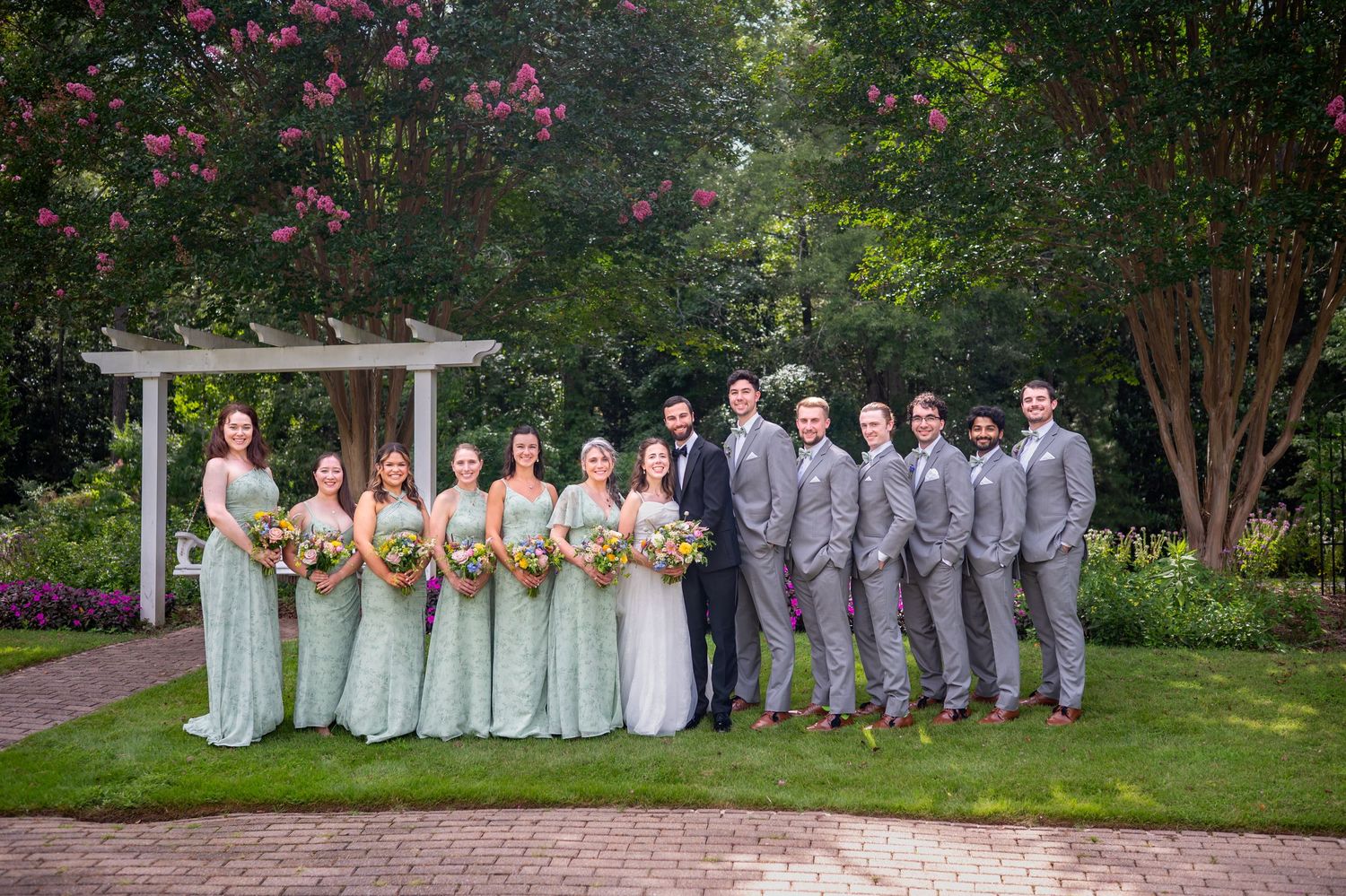 Wedding party in mint green dresses and gray suits pose together in front of garden pergola with blooming flowers.
