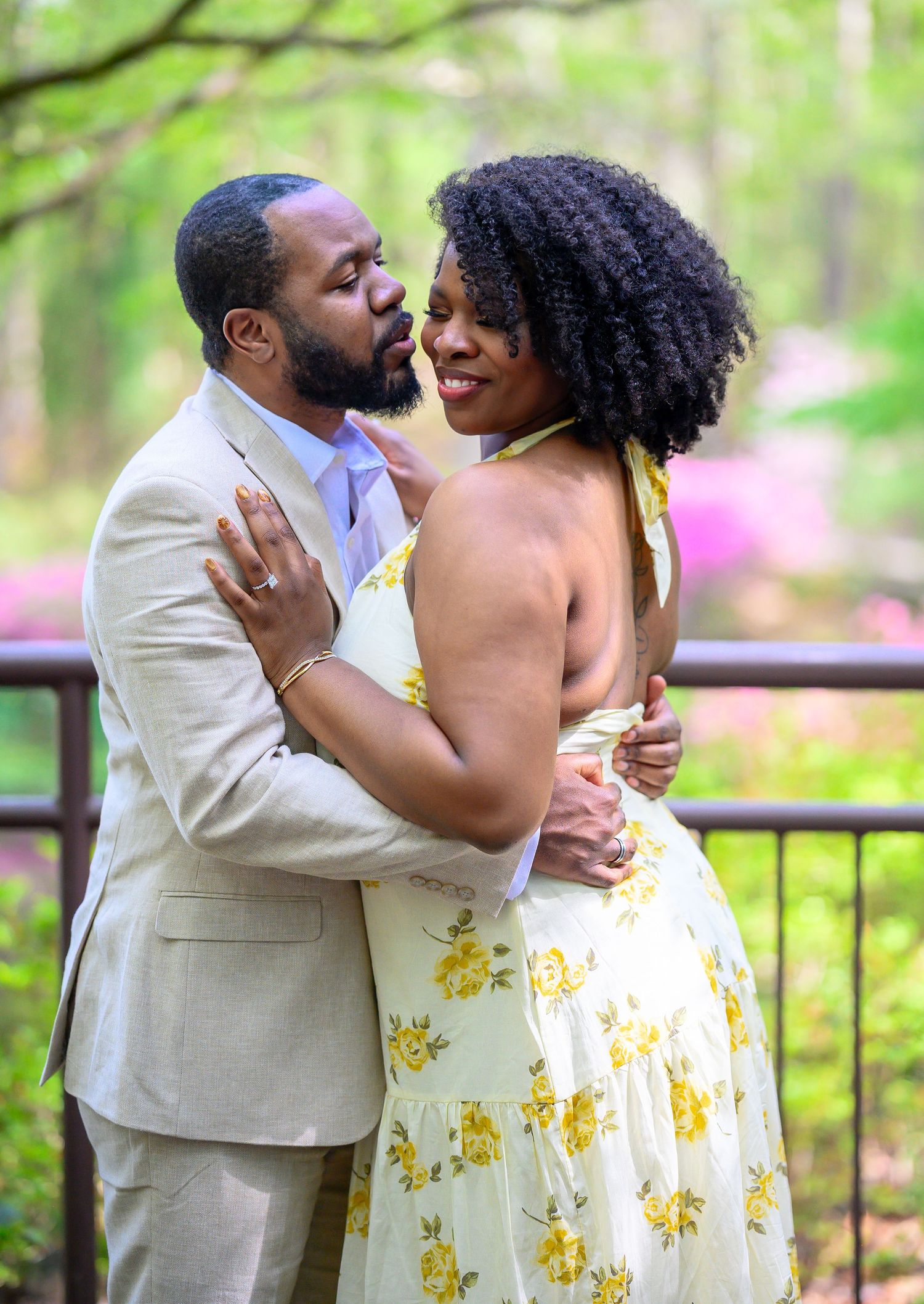 A couple shares a romantic embrace and kiss outdoors with blooming azaleas and green trees in the background.