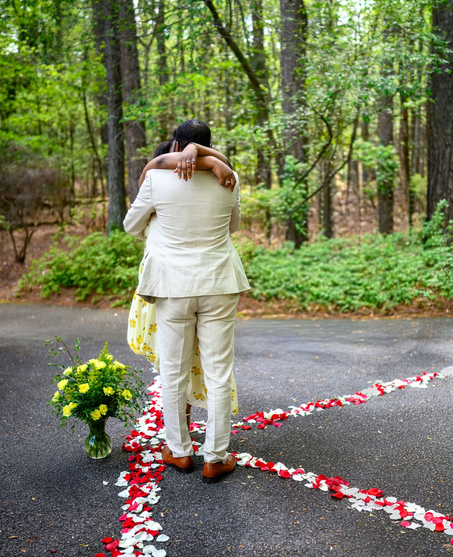 Newly engaged couple embraces beside a heart-shaped rose petal path with yellow flowers in a forest.
