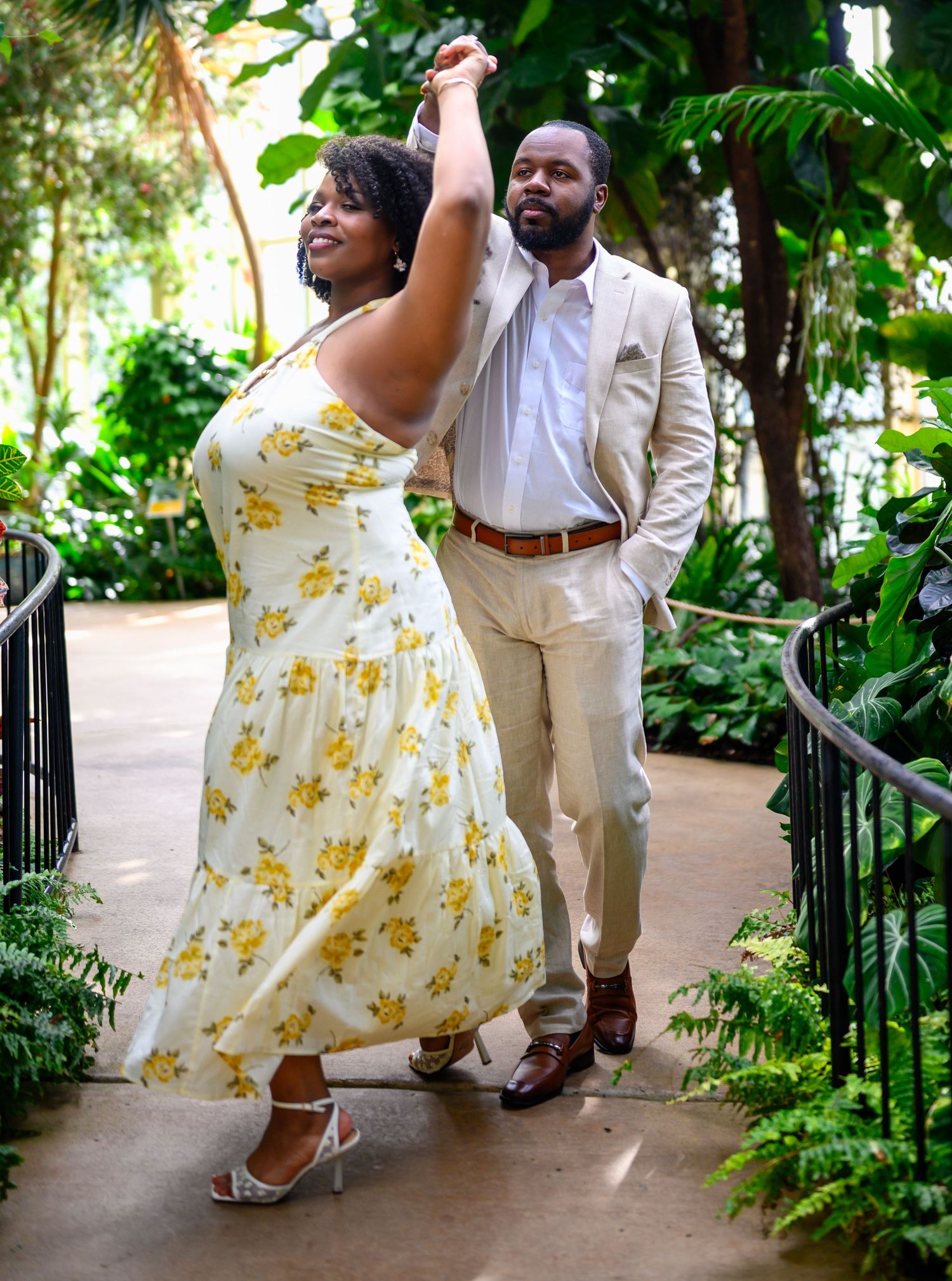 A couple dances joyfully on a garden path, woman in yellow floral dress, man in beige suit.