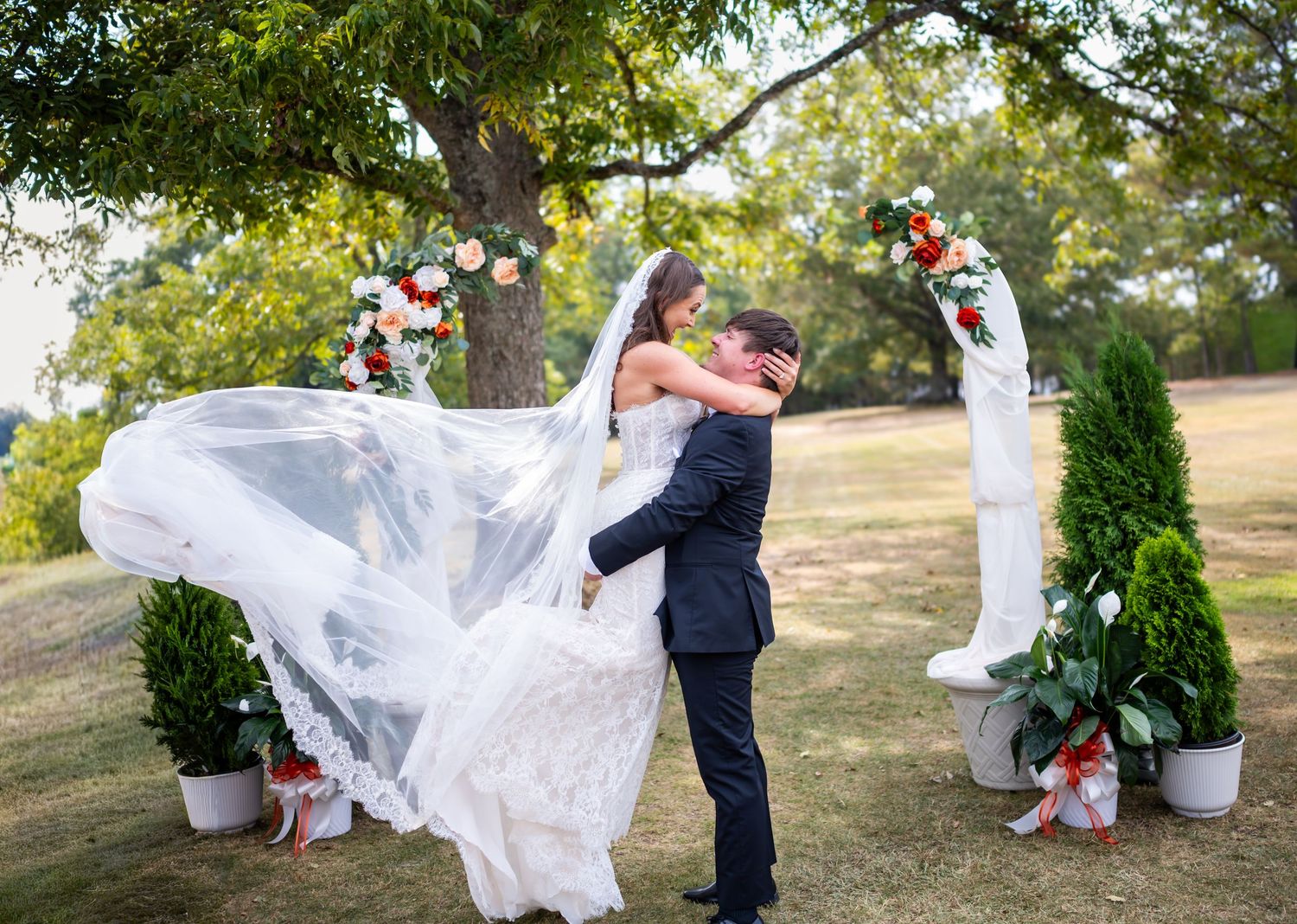 A couple shares a romantic moment outdoors with the veil flowing in the breeze near a decorated wedding arch.