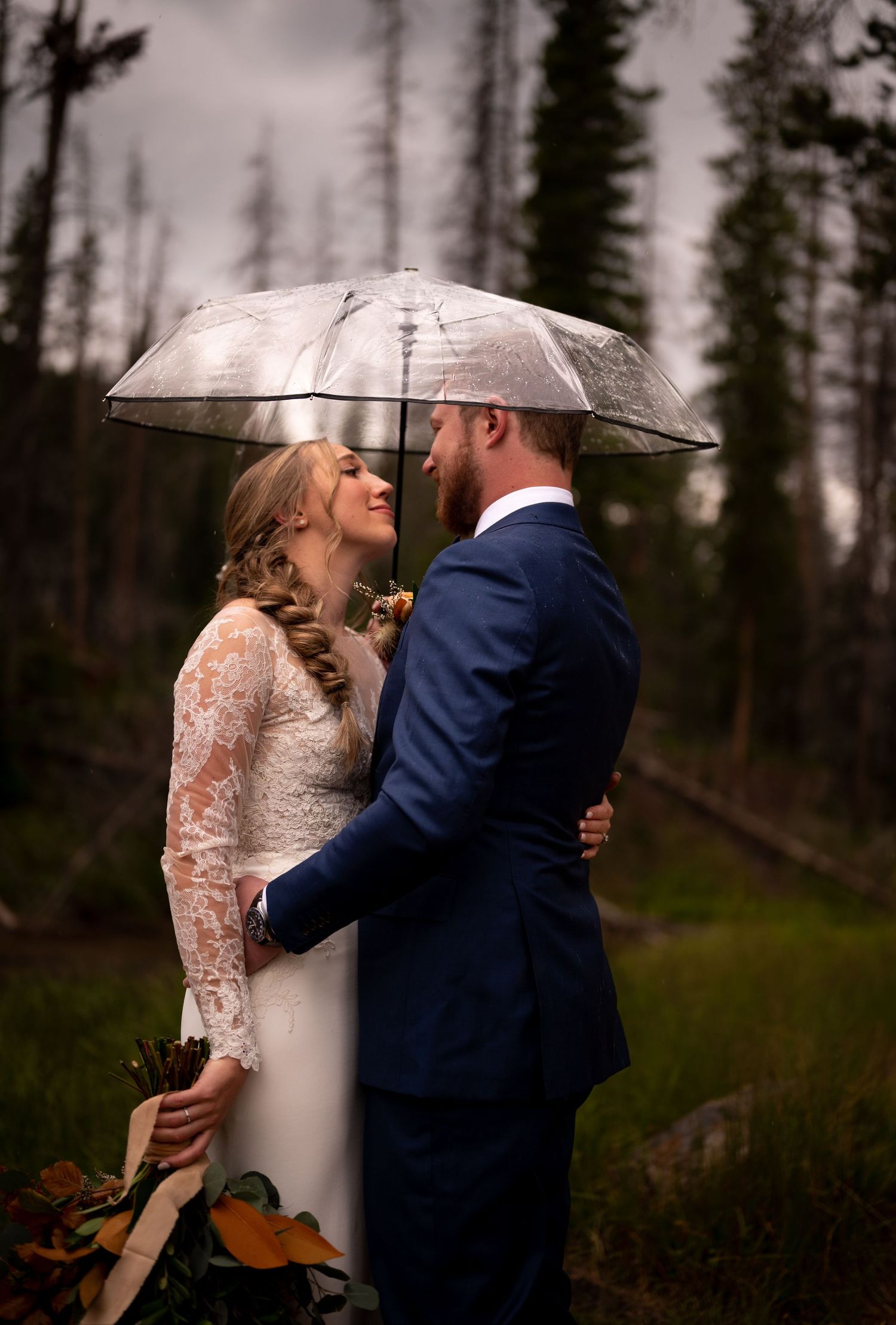 Newlyweds share a romantic moment under clear umbrellas in a forest setting.