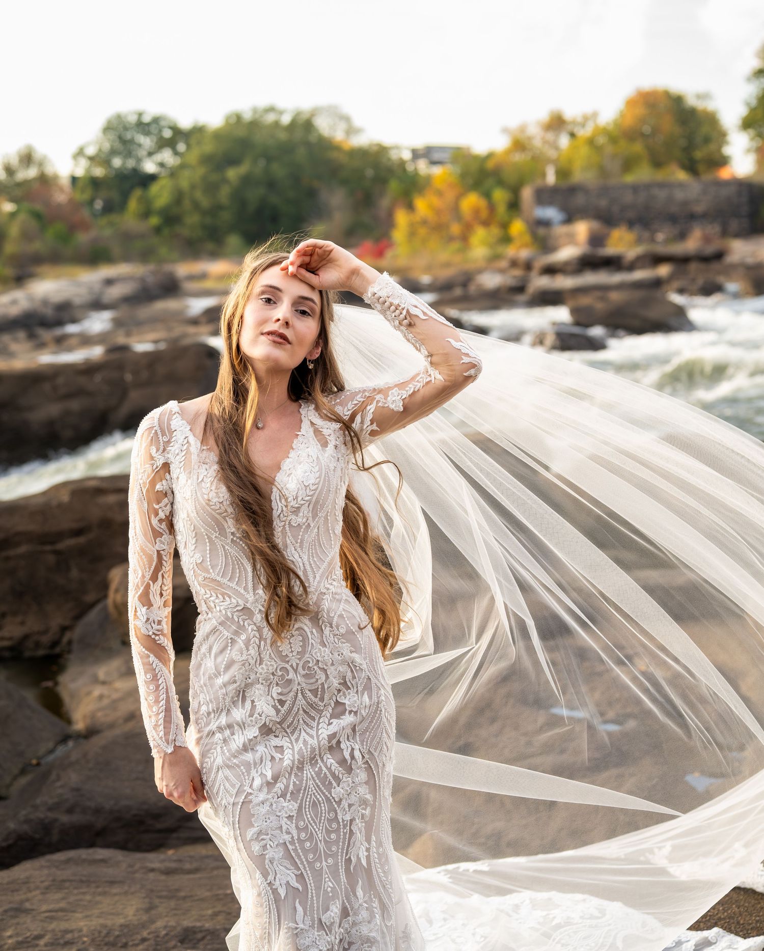Elegant lace wedding dress with flowing veil captured against rocky waterfall backdrop during golden hour.