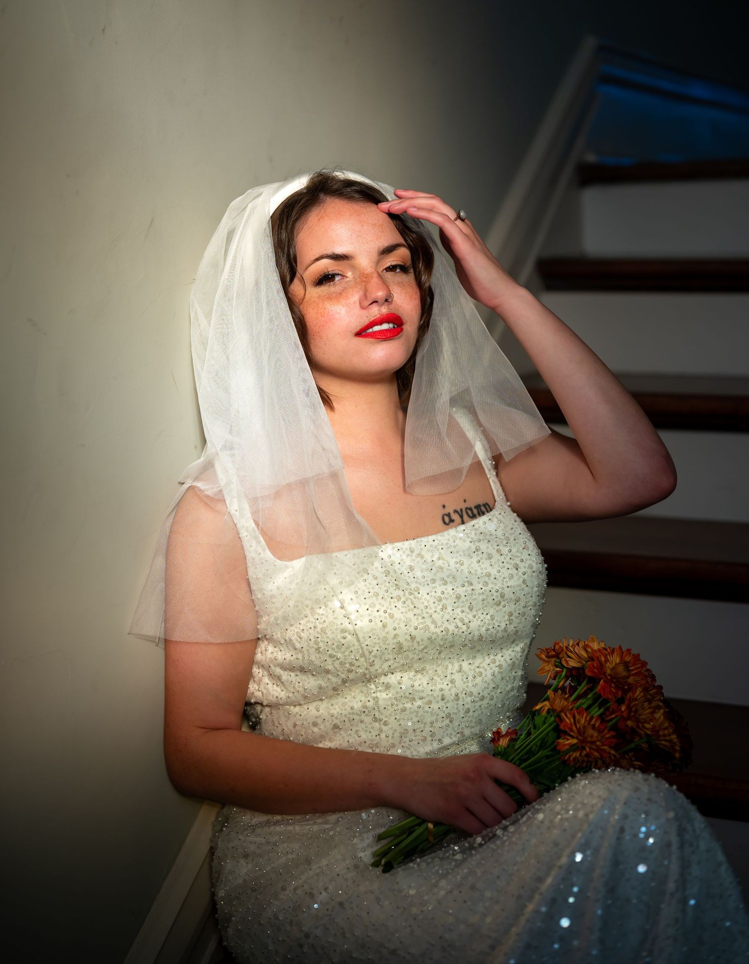 Bride in a sparkling white dress with veil and red lipstick holds an autumn bouquet while posing on stairs.