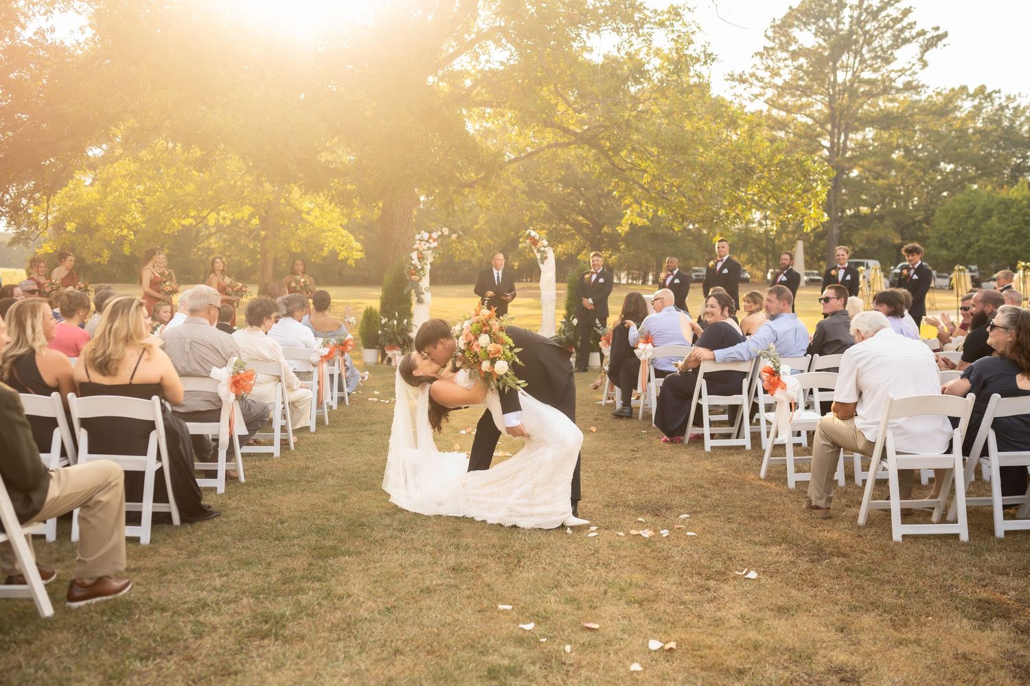 Wedding guests gather outdoors at sunset in white chairs for a garden ceremony celebration.