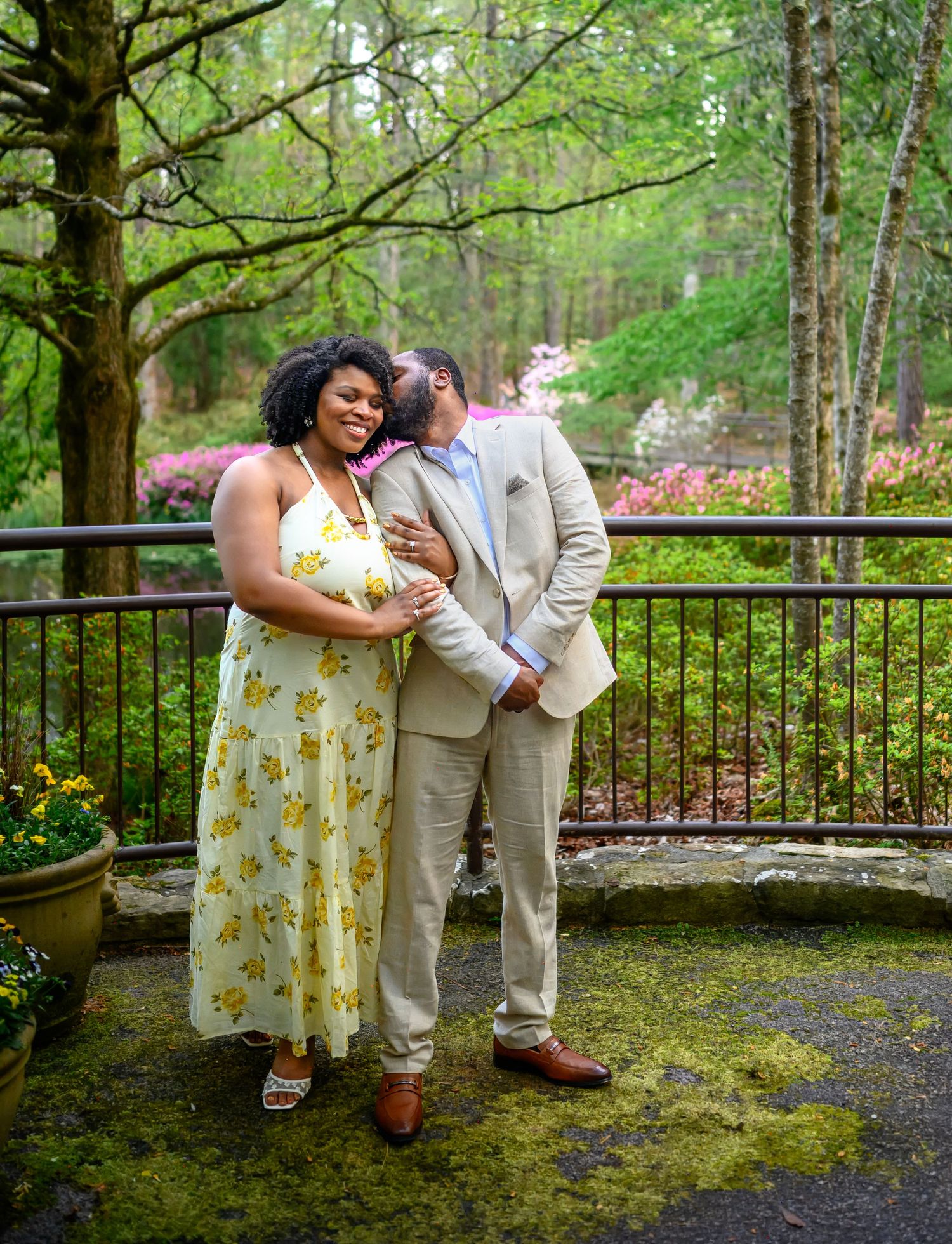 A couple poses affectionately on a mossy terrace surrounded by blooming spring trees and flowers.
