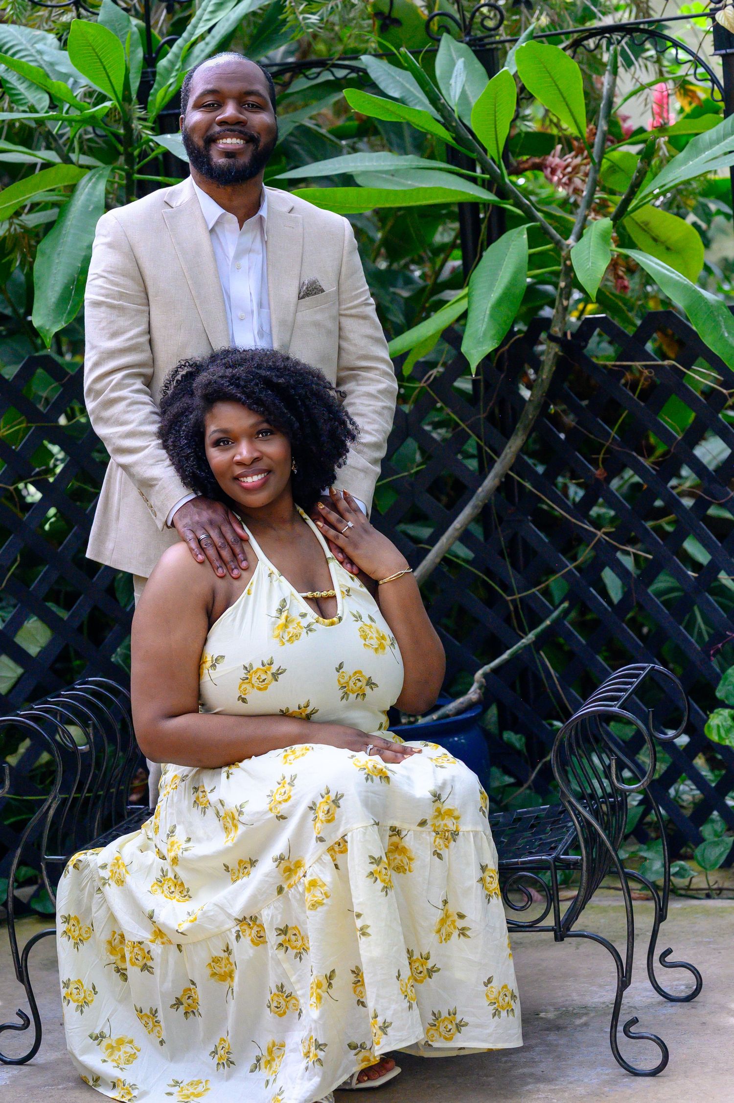 Engaged couple posing in tropical garden, woman seated in yellow floral dress, man standing behind her.