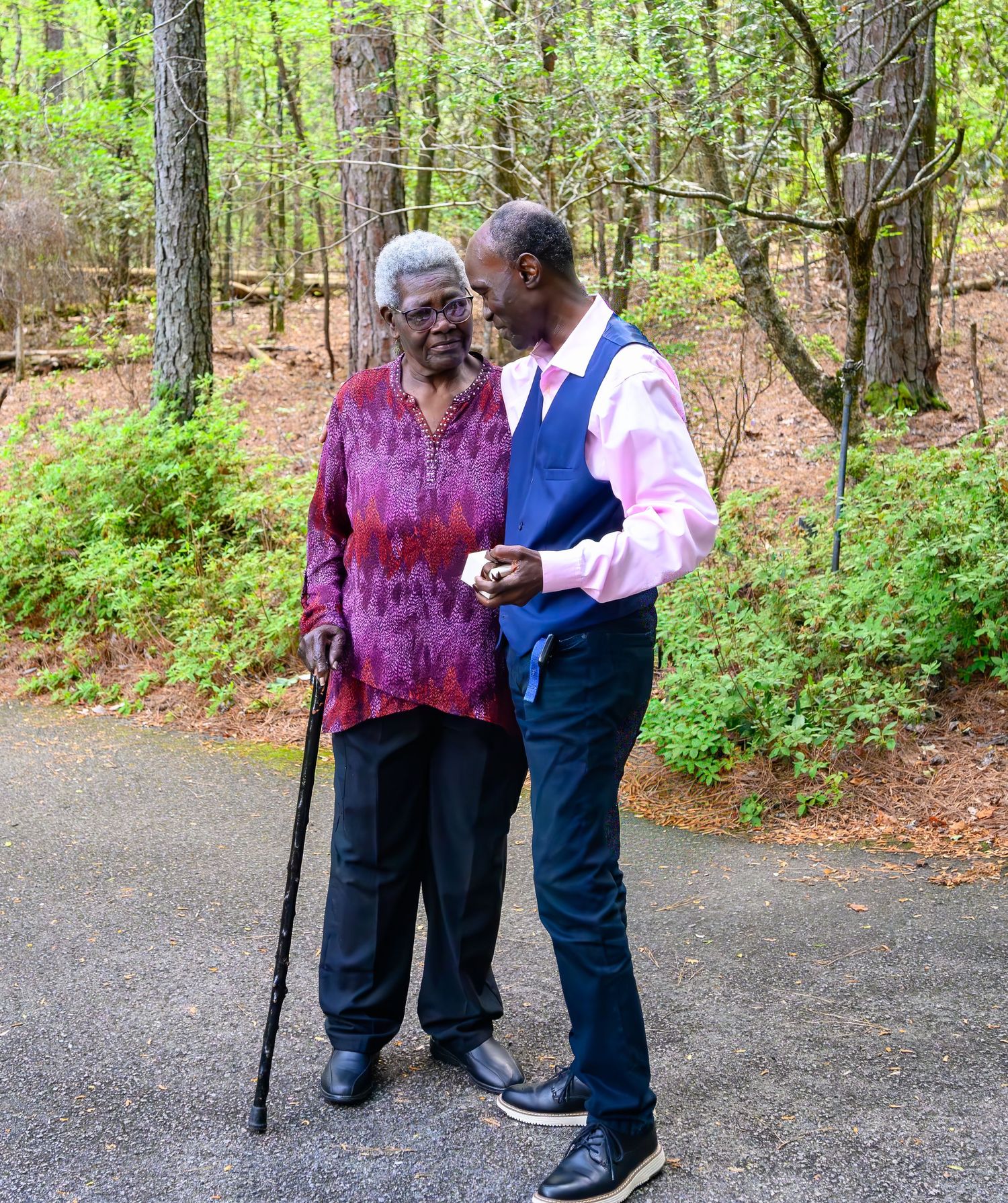 Elderly woman with a cane walks alongside a well-dressed man in a wooded park setting.