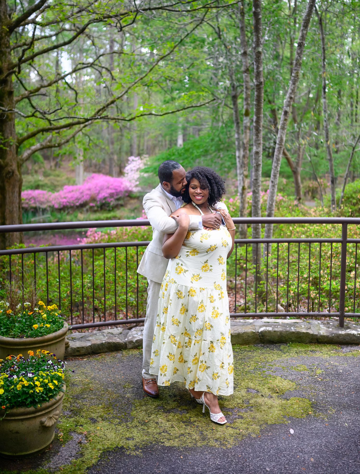 Man in tan suit embraces woman in yellow floral dress on a mossy terrace with blooming azaleas.