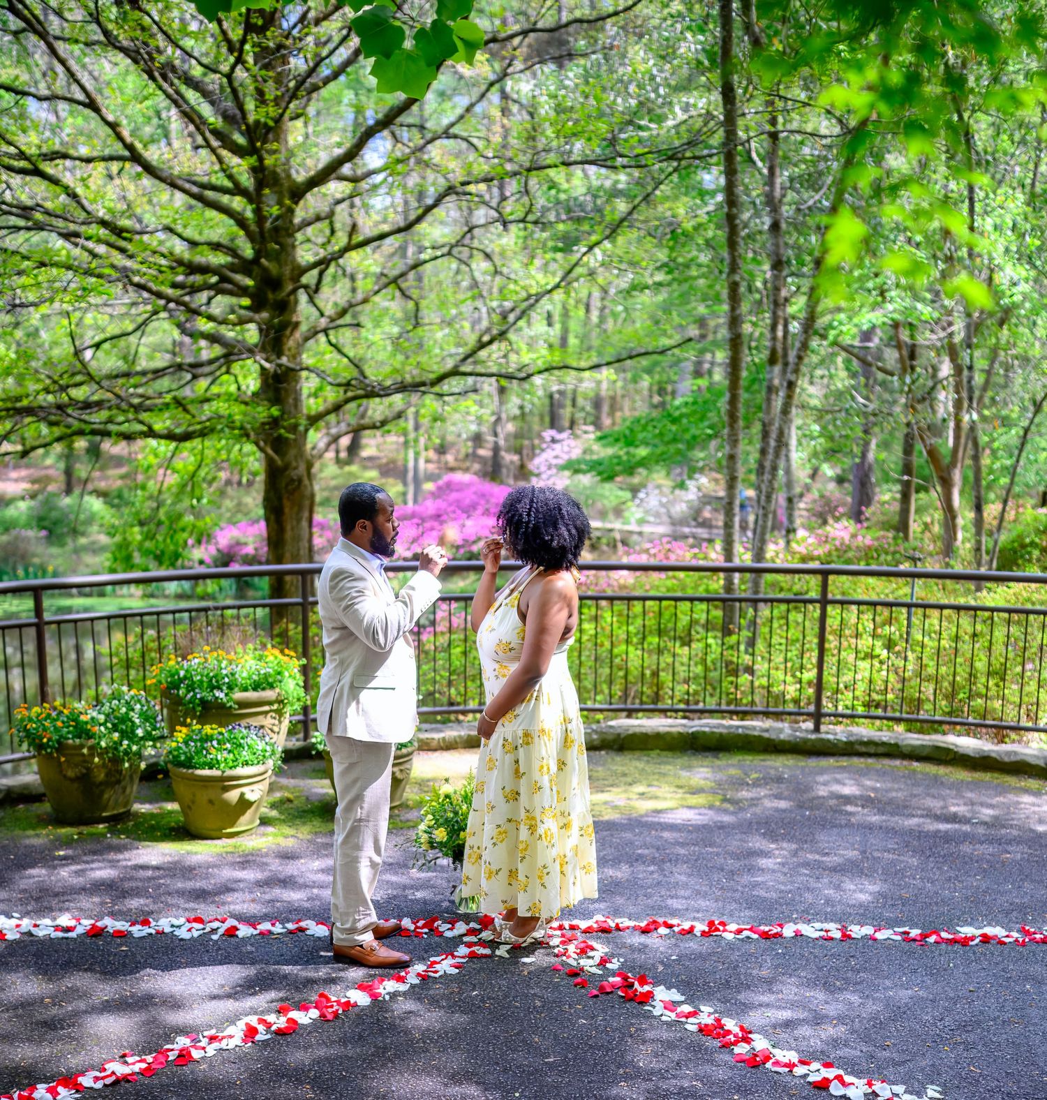 Man kneels to propose to woman in yellow floral dress on a rose petal-decorated garden patio.