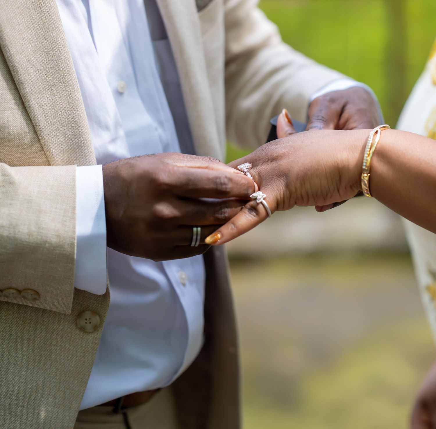 A groom places a diamond ring on his bride's finger during an outdoor wedding ceremony, both wearing elegant attire.