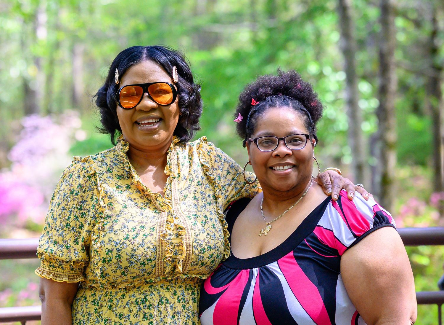 Two women smile together outdoors in a lush green garden with colorful azaleas in the background.