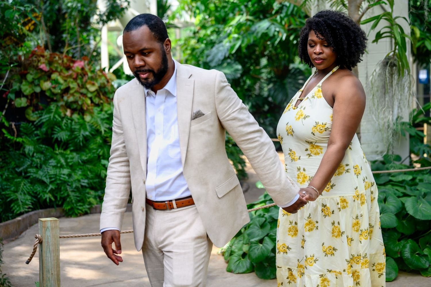 An engaged couple walks hand in hand along a wooden pathway surrounded by lush tropical greenery.