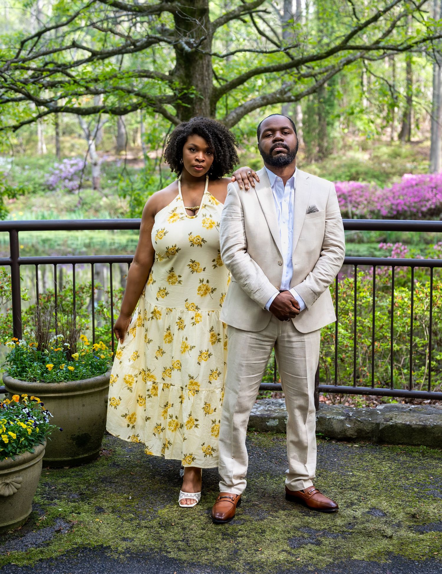 Couple standing together on a garden terrace with blooming azaleas and a large tree in the background.