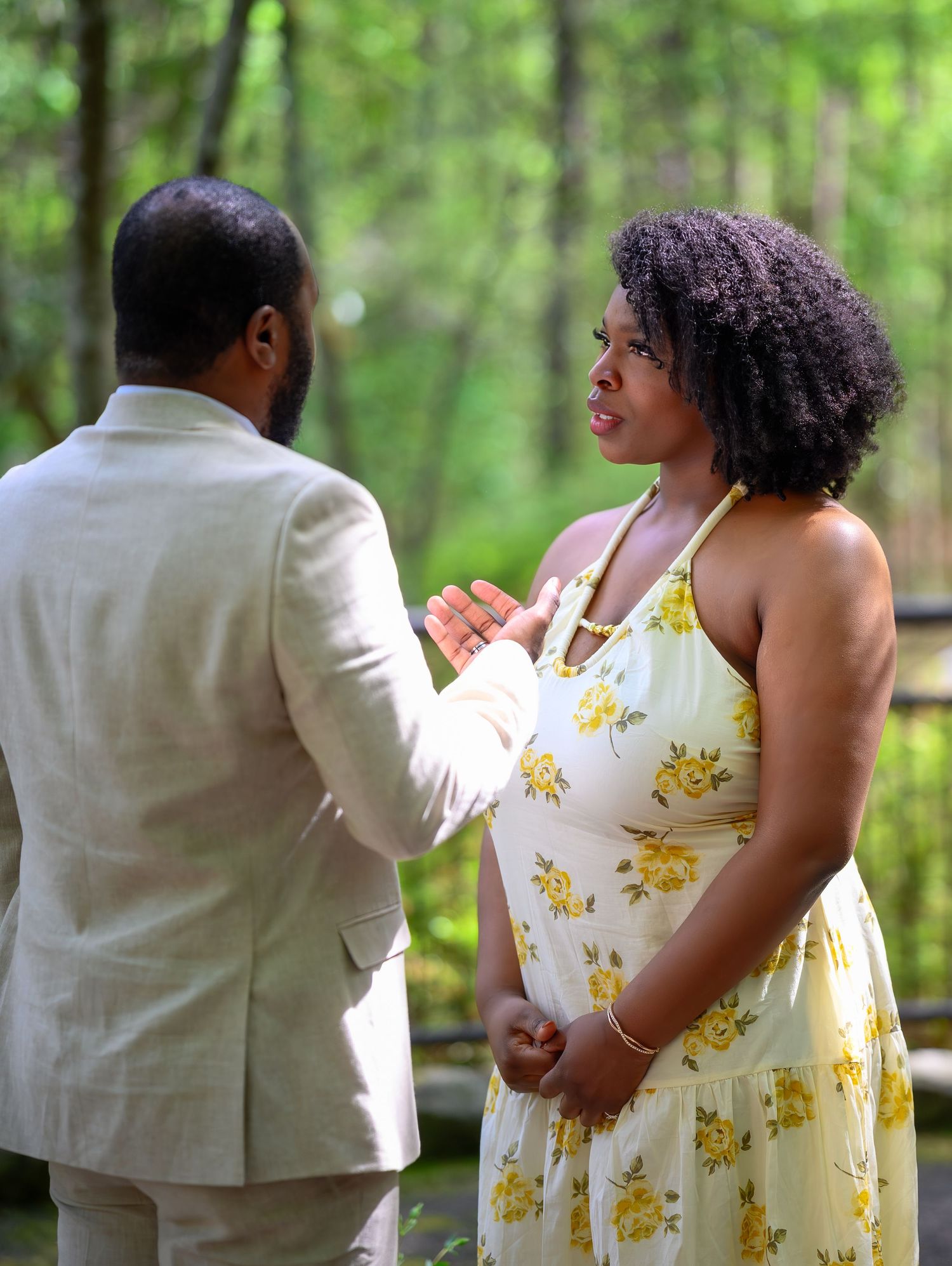 A pregnant woman in a yellow floral dress shares a tender moment with a man in a beige suit outdoors.