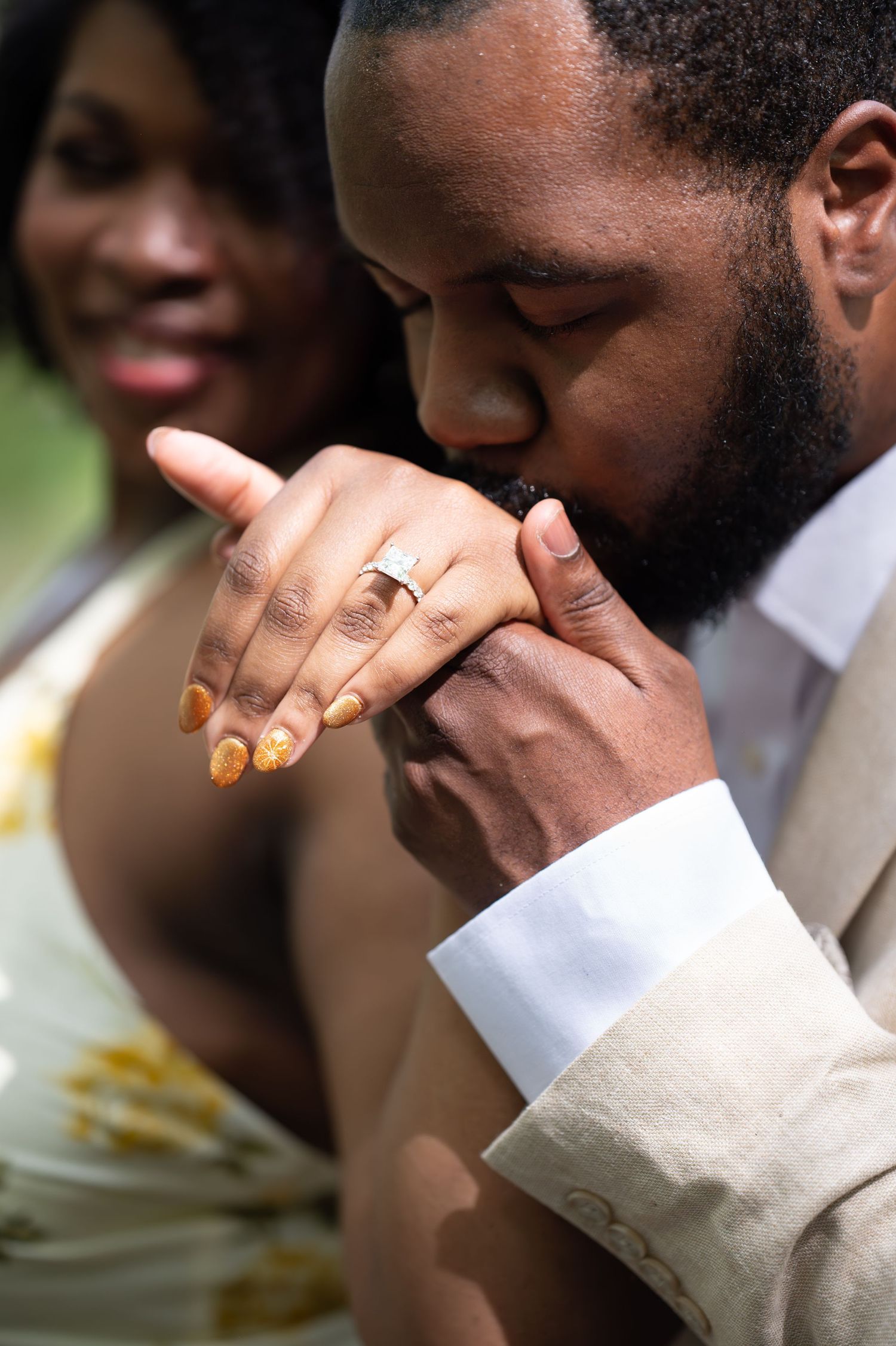 A bearded man tenderly kisses his fiancees hand adorned with a diamond engagement ring as she smiles behind him.