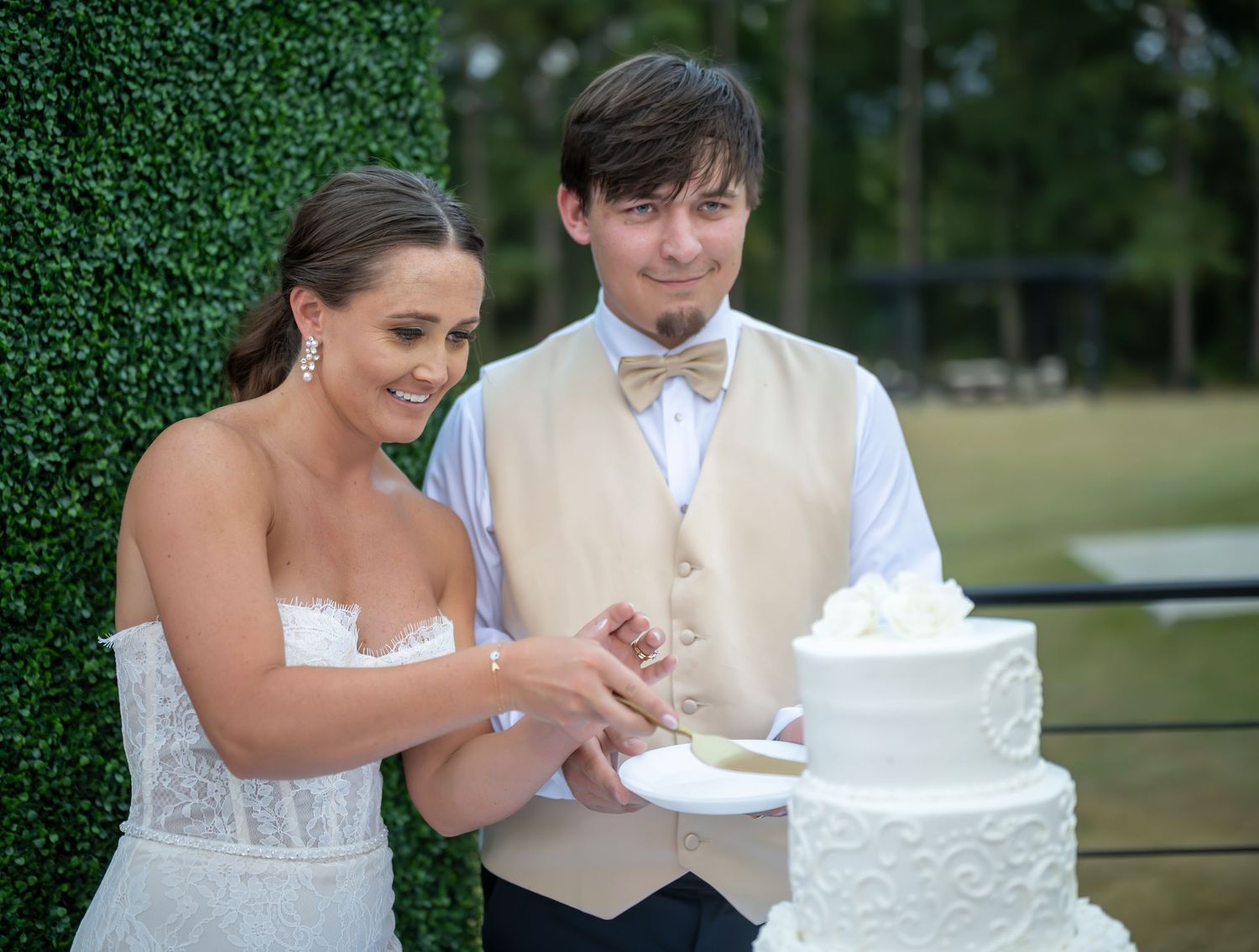 Newlyweds cut their elegant white tiered wedding cake together at an outdoor reception.