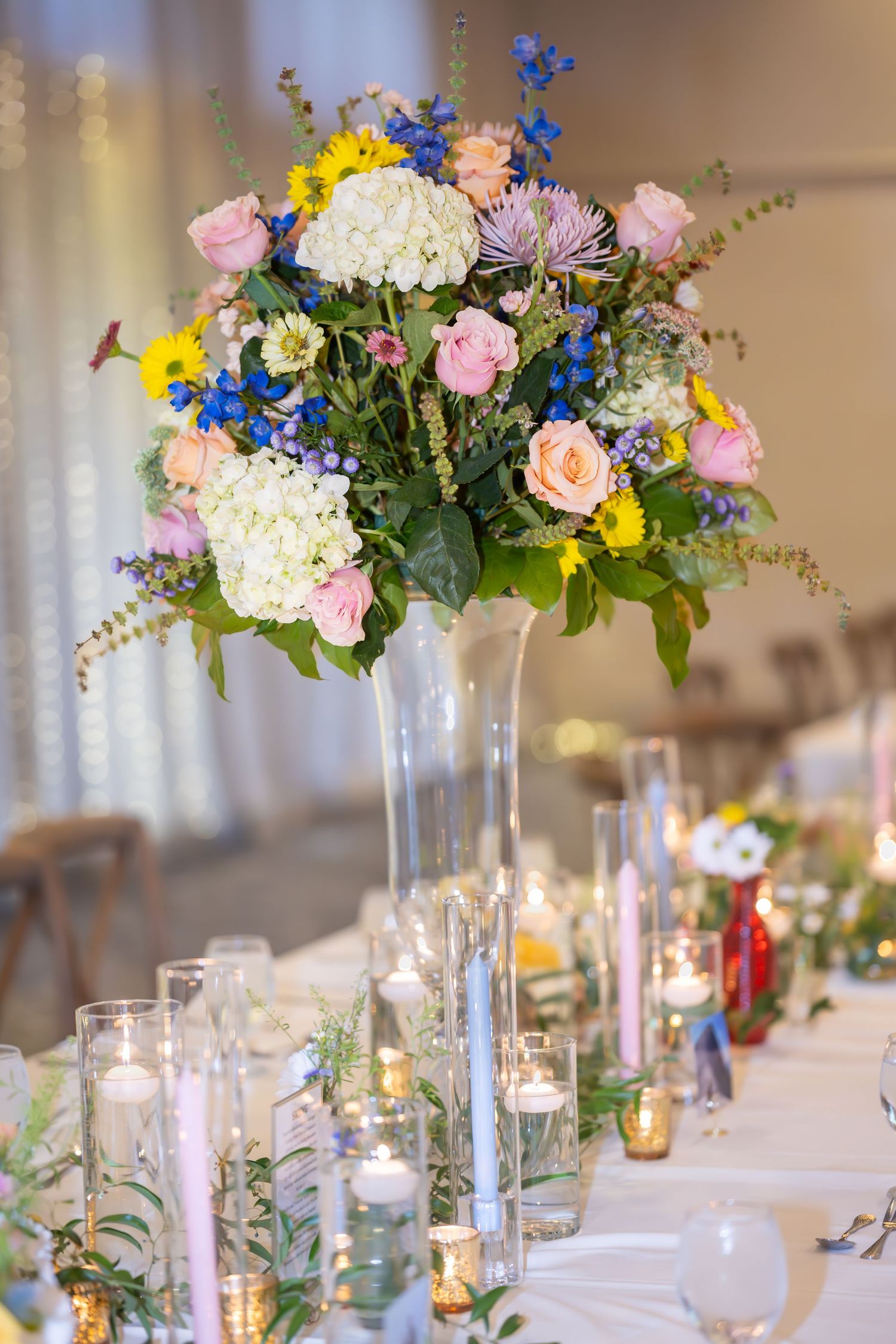 Elegant wedding reception tables decorated with tall glass vases filled with pink, blue, and yellow floral centerpieces.