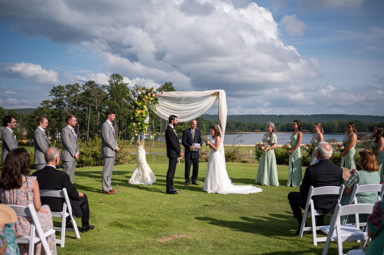 Panoramic view of lakeside wedding ceremony with white chairs and floral arch decorations.