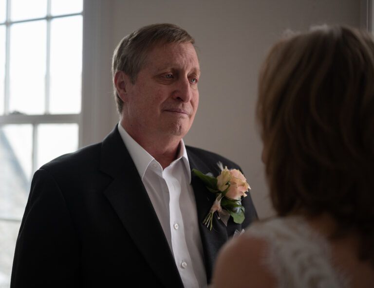 Wedding couple share intimate moment in room with large windows and natural lighting.