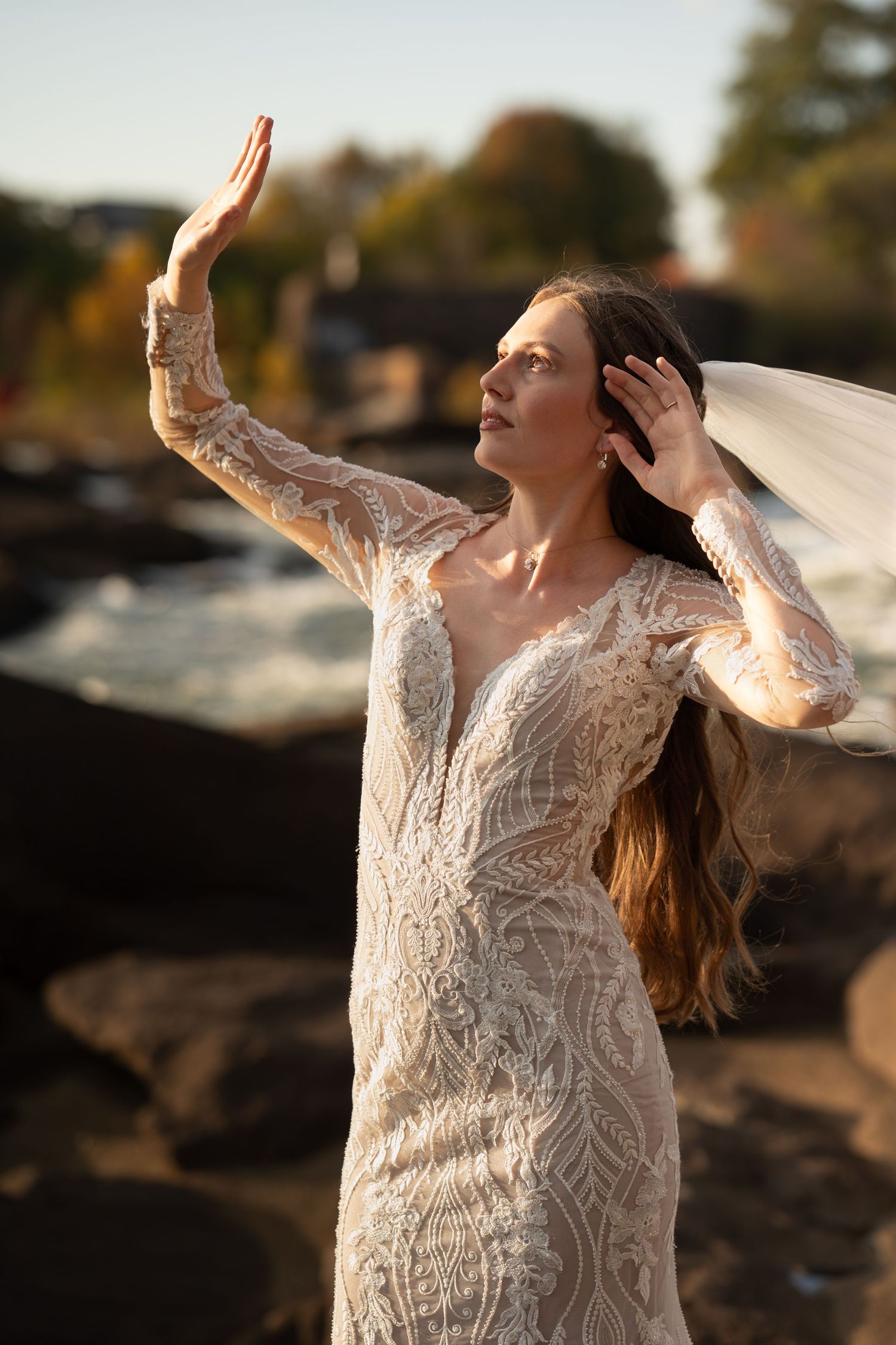 Ethereal bridal pose with arms raised as veil catches sunlight against rocky landscape.