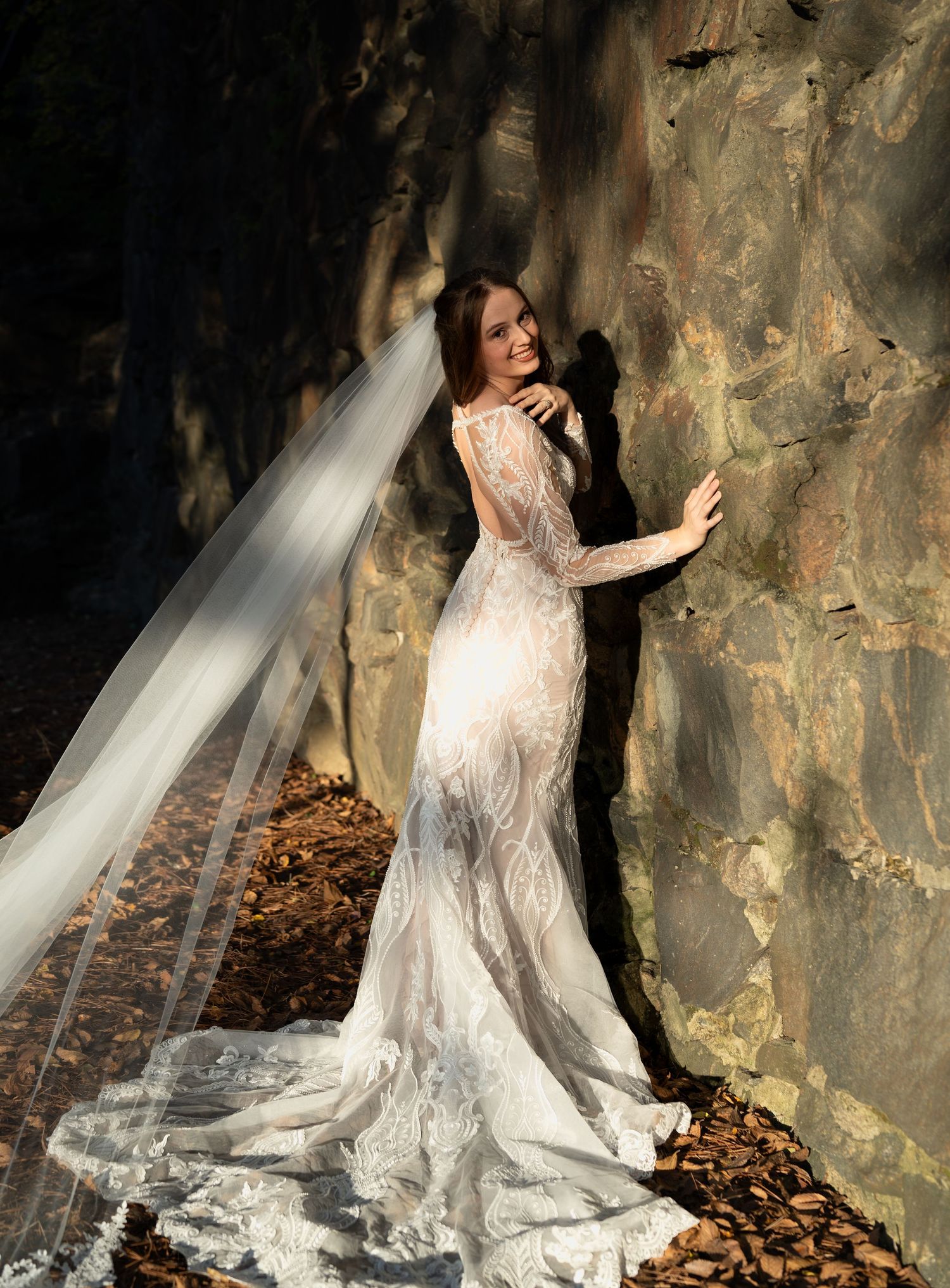 A bride in a lace wedding dress poses against a stone wall with autumn leaves and flowing veil in dramatic lighting.