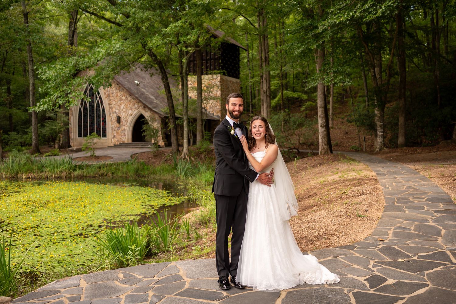 Bride and groom walking hand in hand on stone path with chapel and forest in background.