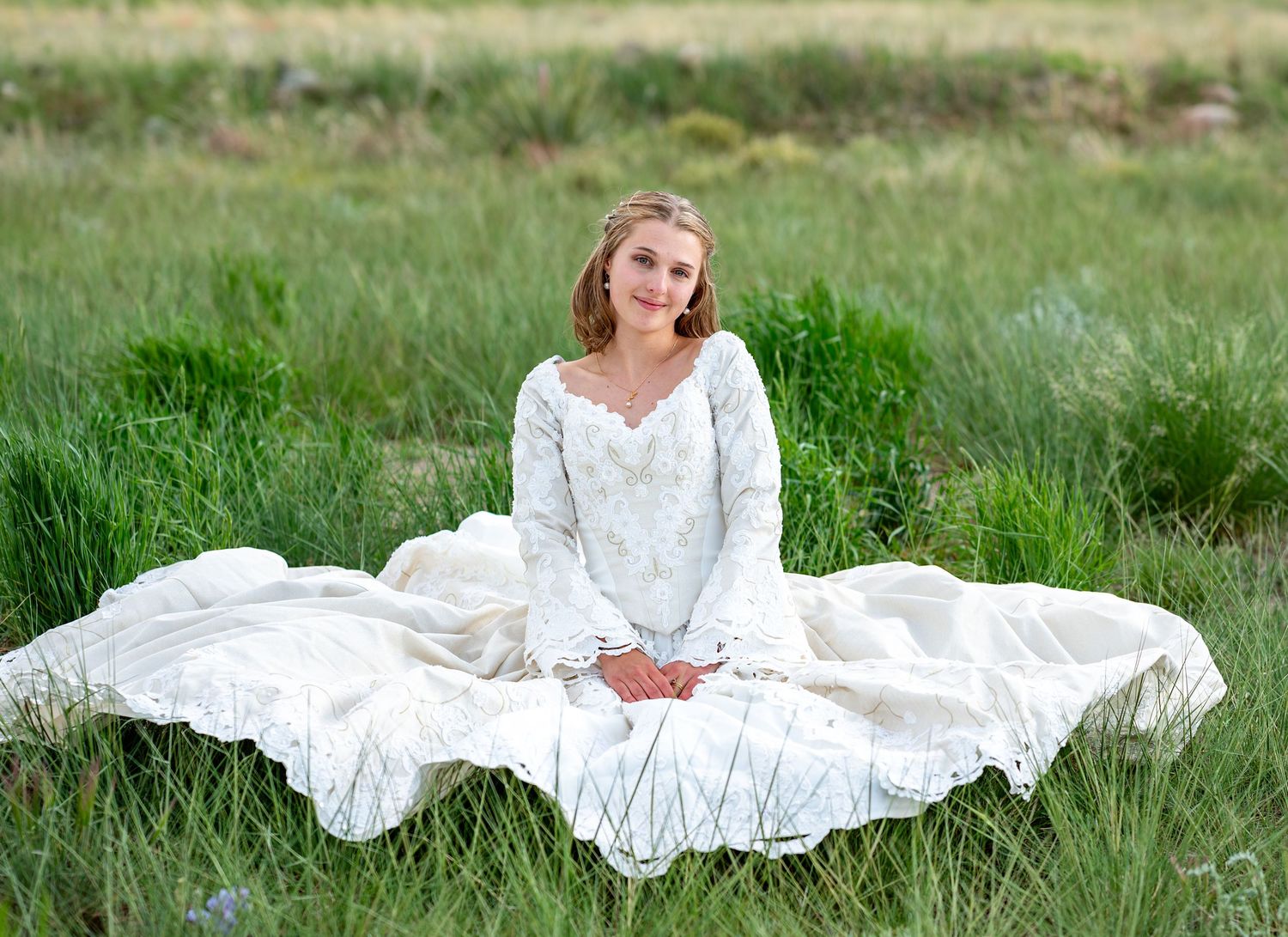 Woman in white lace wedding dress posing gracefully in a green meadow with mountain views.
