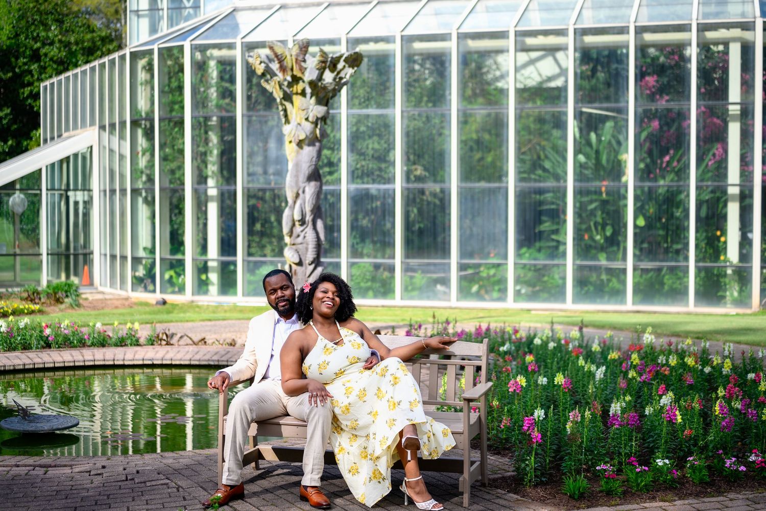 Couple relaxing on a bench beside a pond in front of a botanical greenhouse.