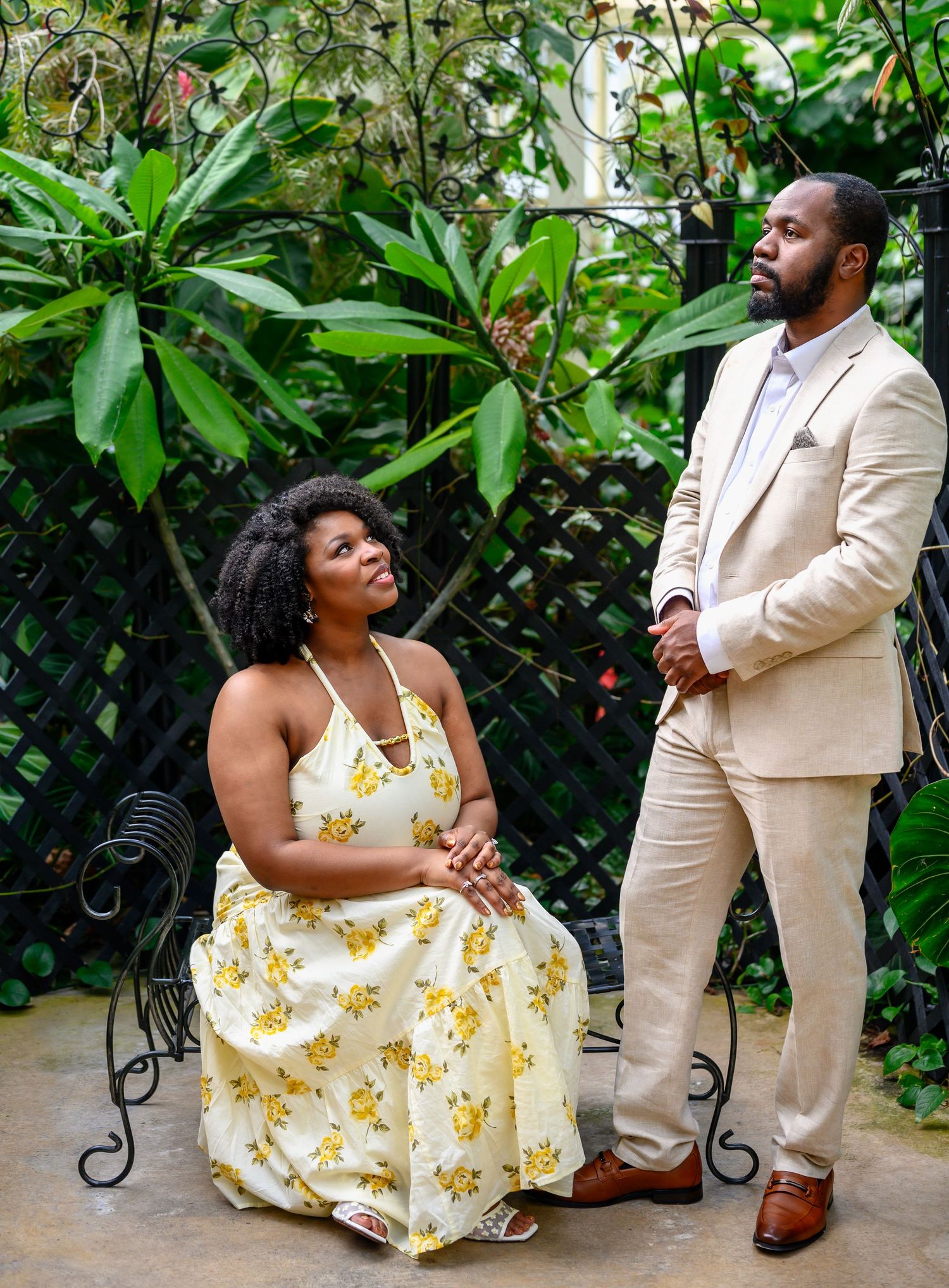 Woman seated in a garden chair gazing up at a man in a beige suit among lush tropical plants.