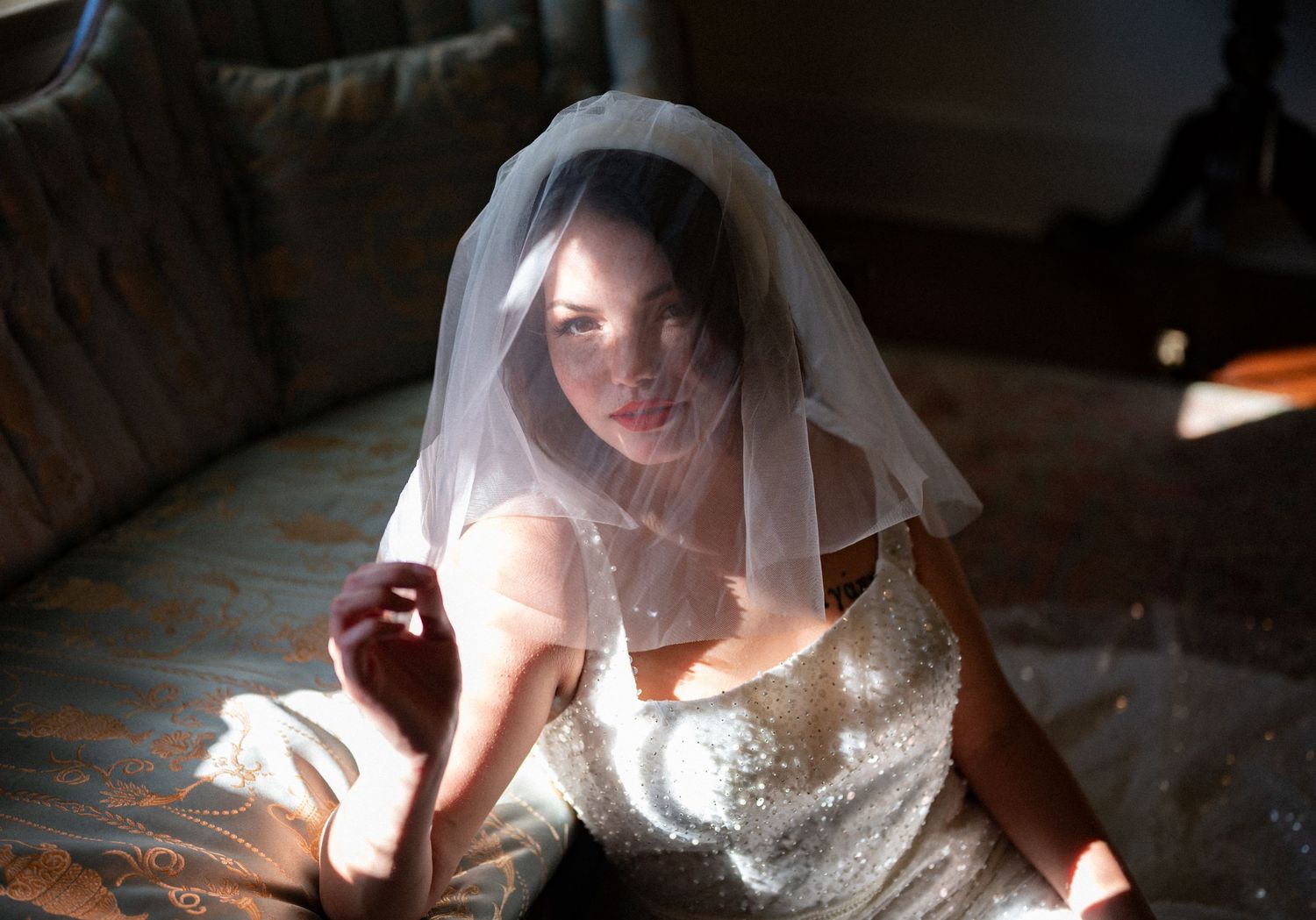 A bride in a white veil and sparkly dress sits on a couch in soft natural lighting.