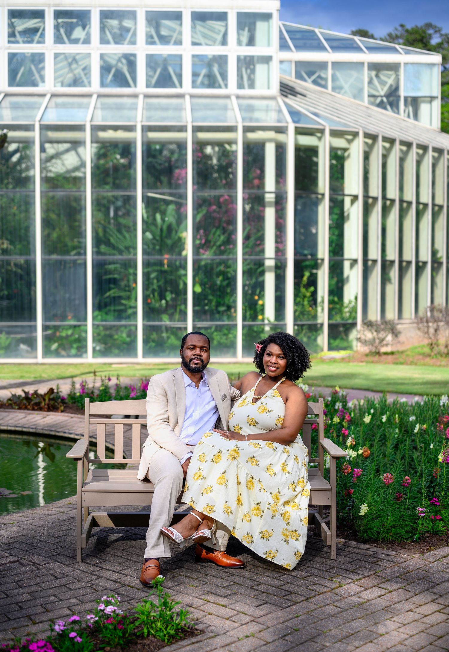 Couple posing on a wooden bench in front of a large glass greenhouse surrounded by colorful flowers.