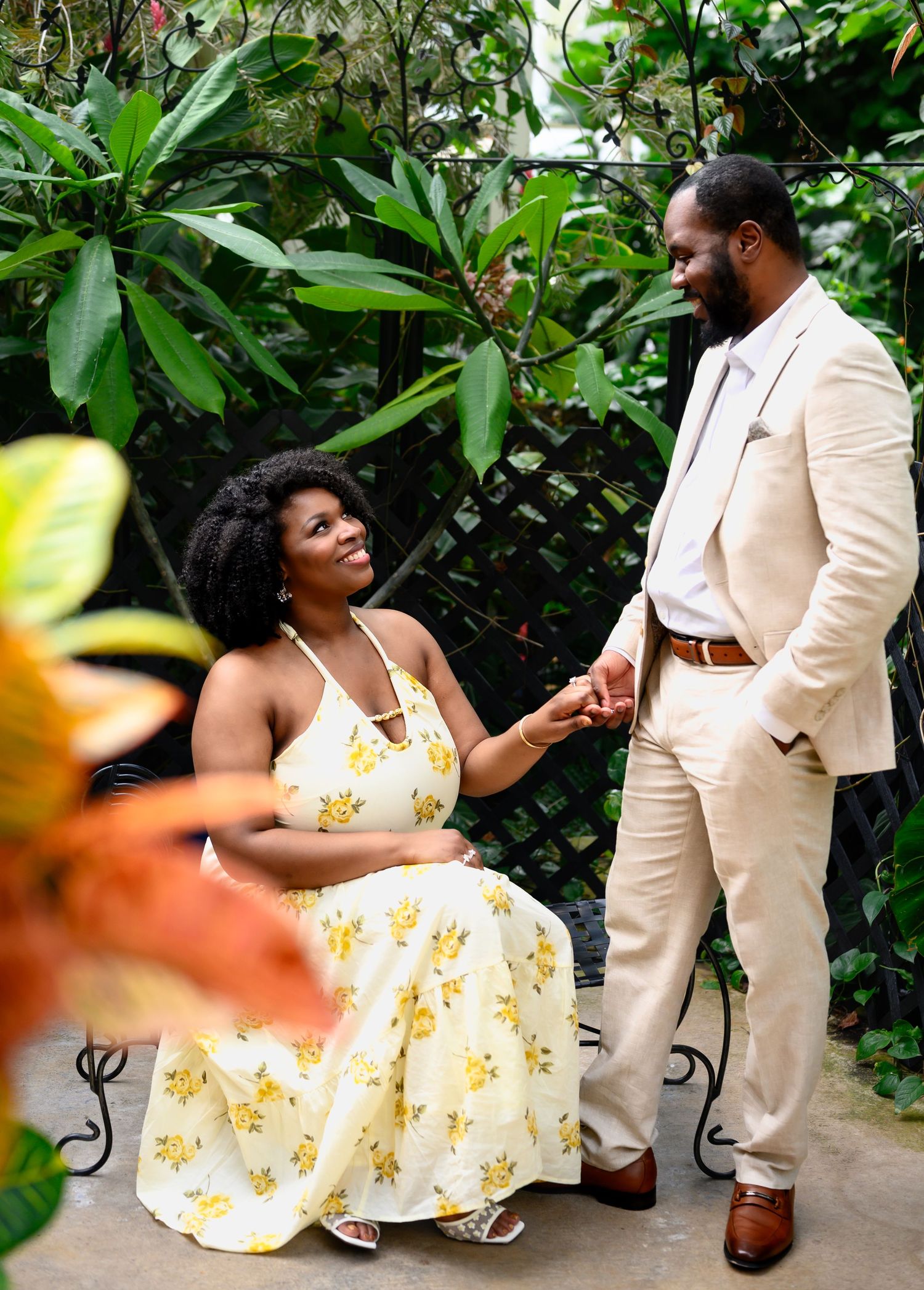 Engaged couple holding hands and smiling at each other in lush green tropical garden setting.