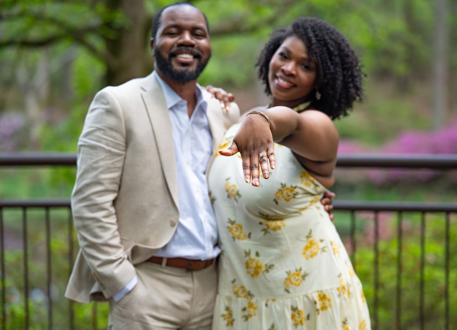 Smiling newly engaged Black couple poses together outdoors, woman showing off engagement ring in garden.