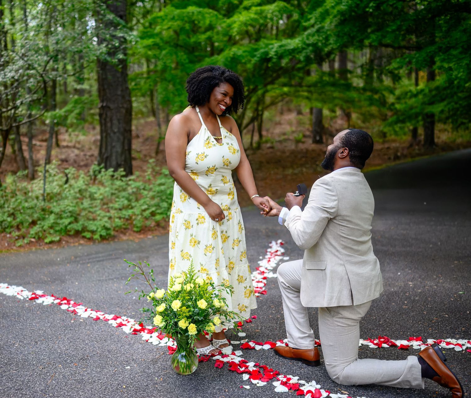 Couple sharing joyful marriage proposal moment on rose petal lined path surrounded by lush green woodland trees.