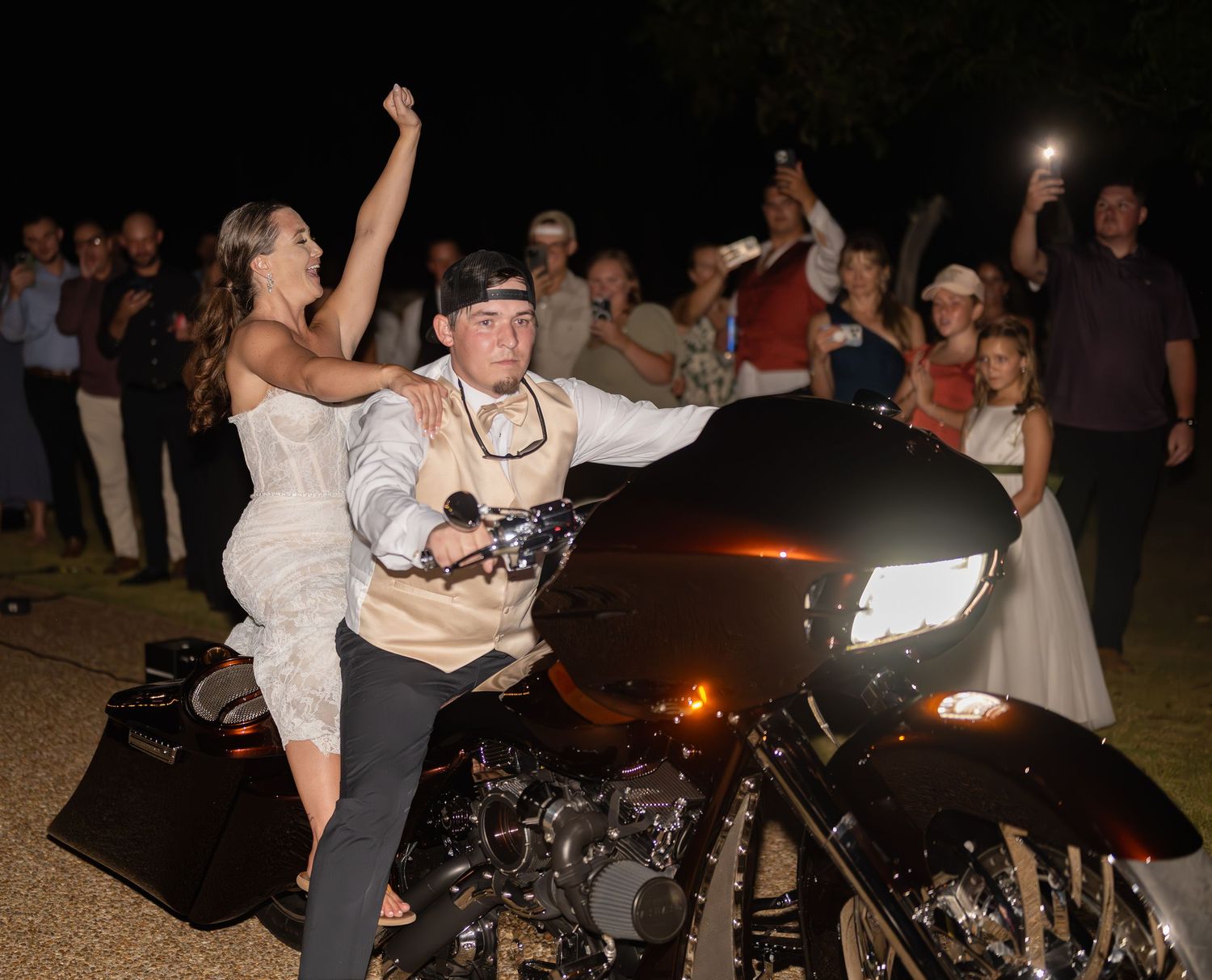 Newlyweds pose excitedly on motorcycle during wedding reception exit.