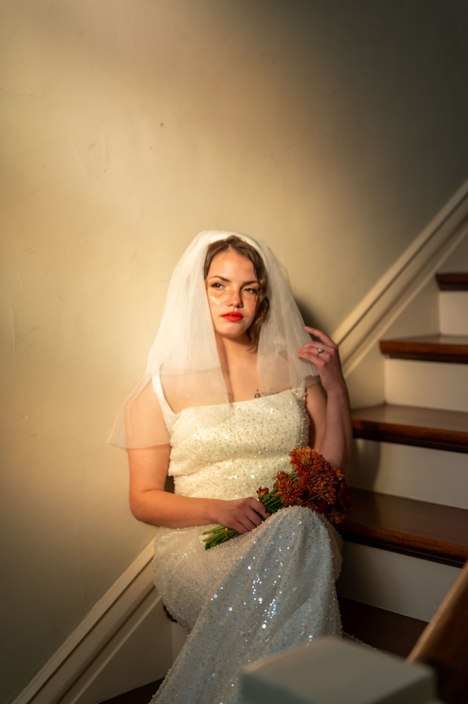 A bride in a sparkling white wedding dress holding flowers stands near indoor stairs in soft lighting.