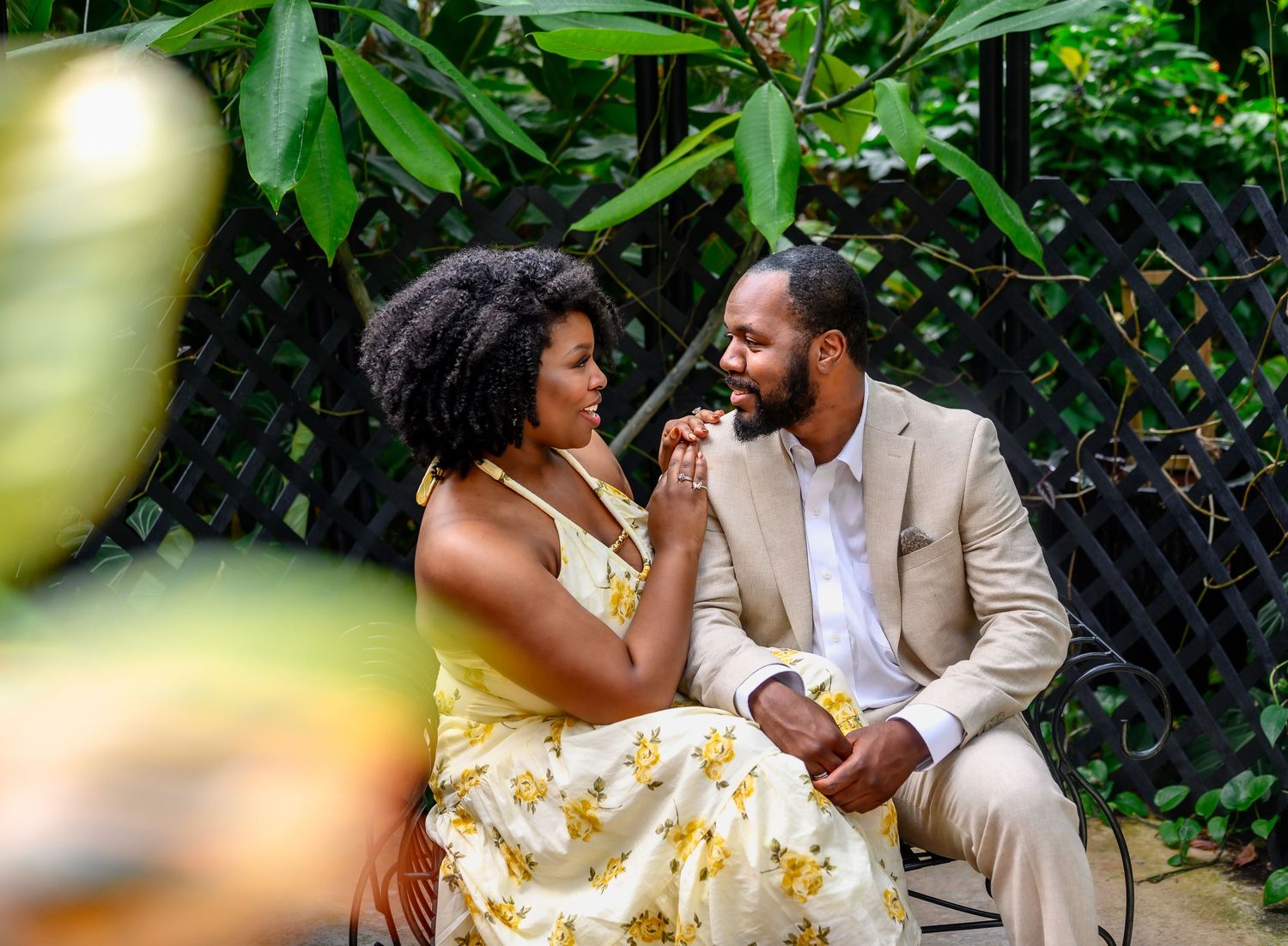 An engaged couple sits together in a garden as the woman kisses the mans hand while he gazes at her lovingly.