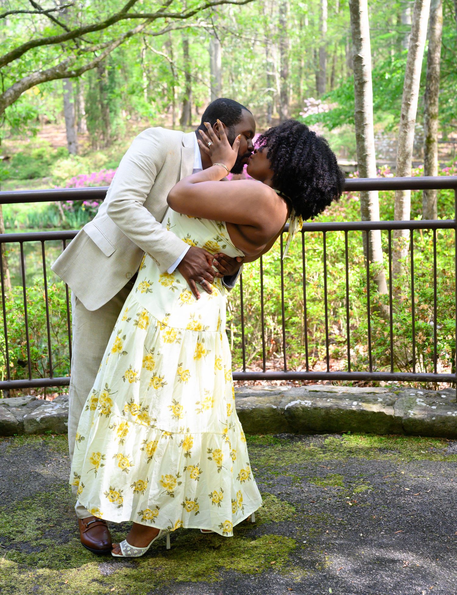 A couple shares a romantic dip kiss on a stone terrace surrounded by a wooded garden.