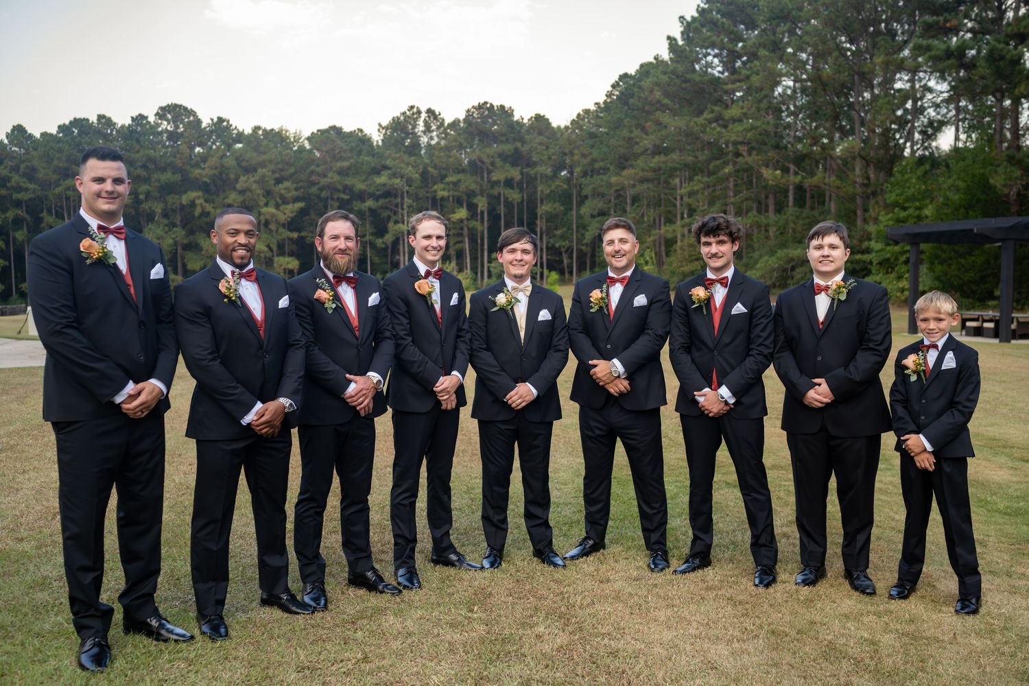 Groomsmen in black tuxedos line up for formal wedding photo outdoors.