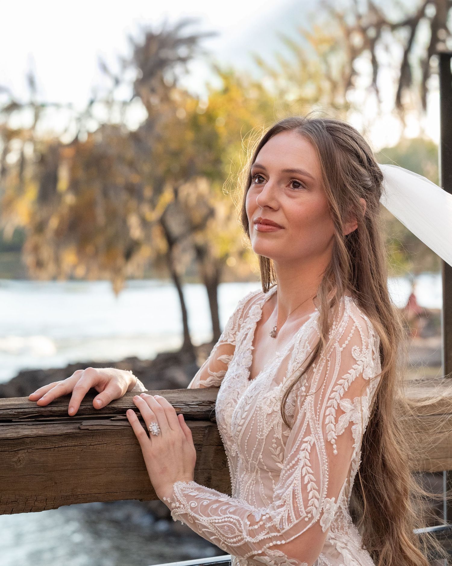 Bride in lace long sleeve wedding dress poses thoughtfully by wooden railing at sunset near water.