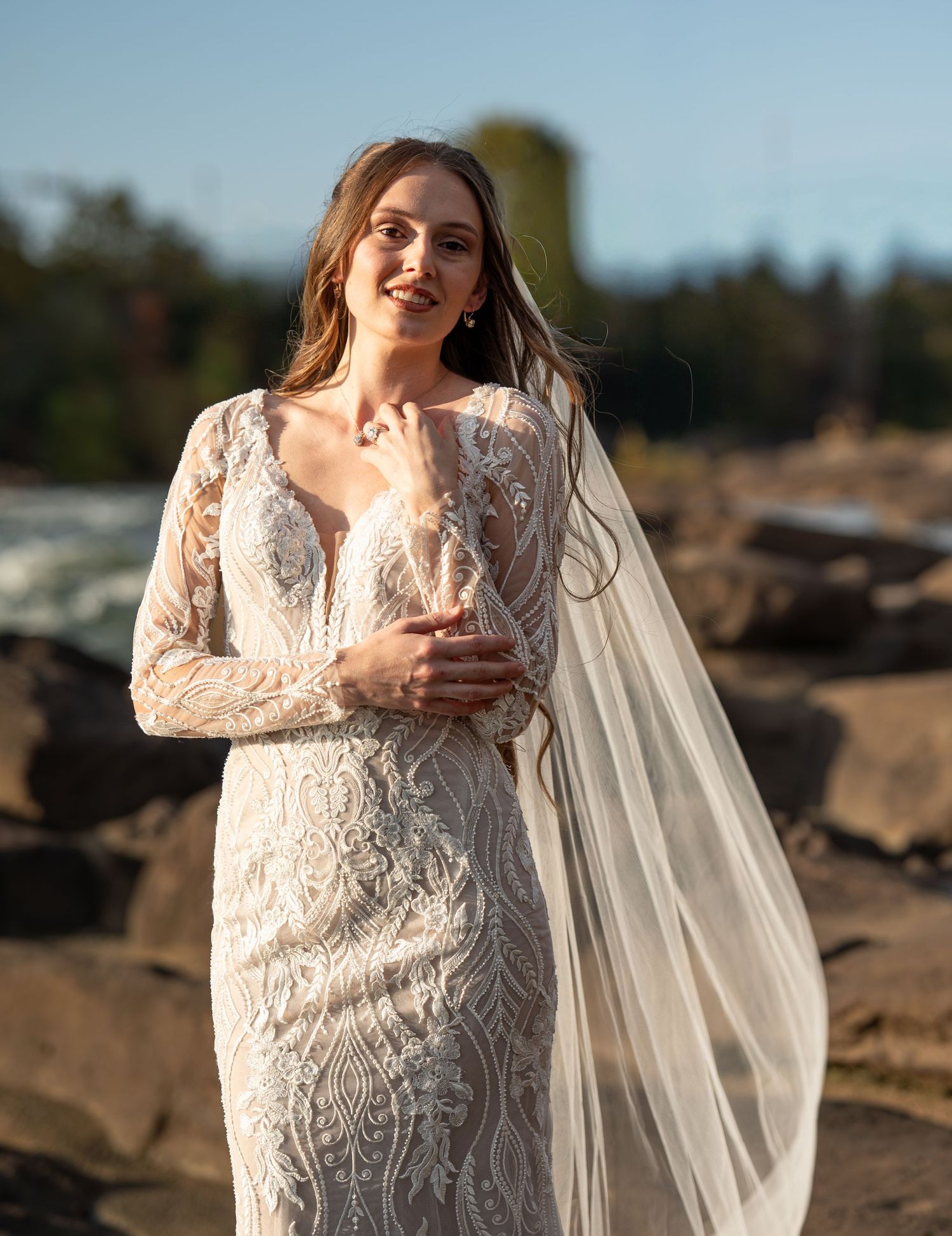 A bride in a lace wedding dress with long sleeves and flowing veil poses on rocky terrain at sunset.