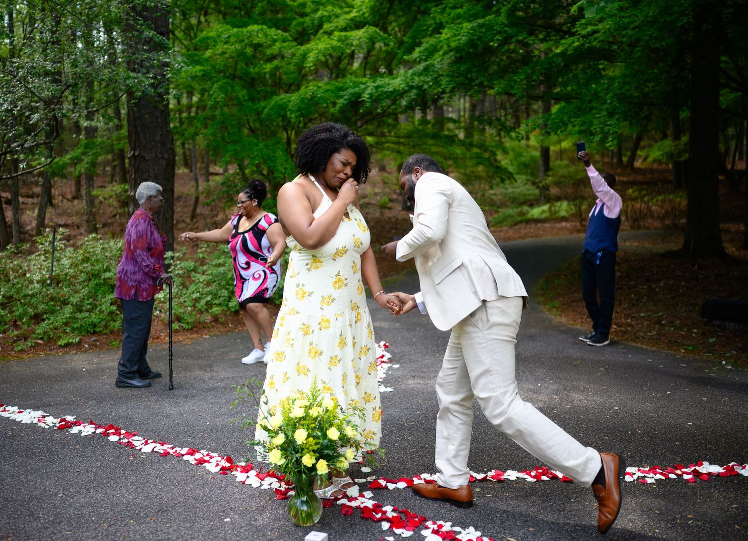 Couple sharing a joyful moment on a petal-lined path in a wooded setting during a surprise proposal.