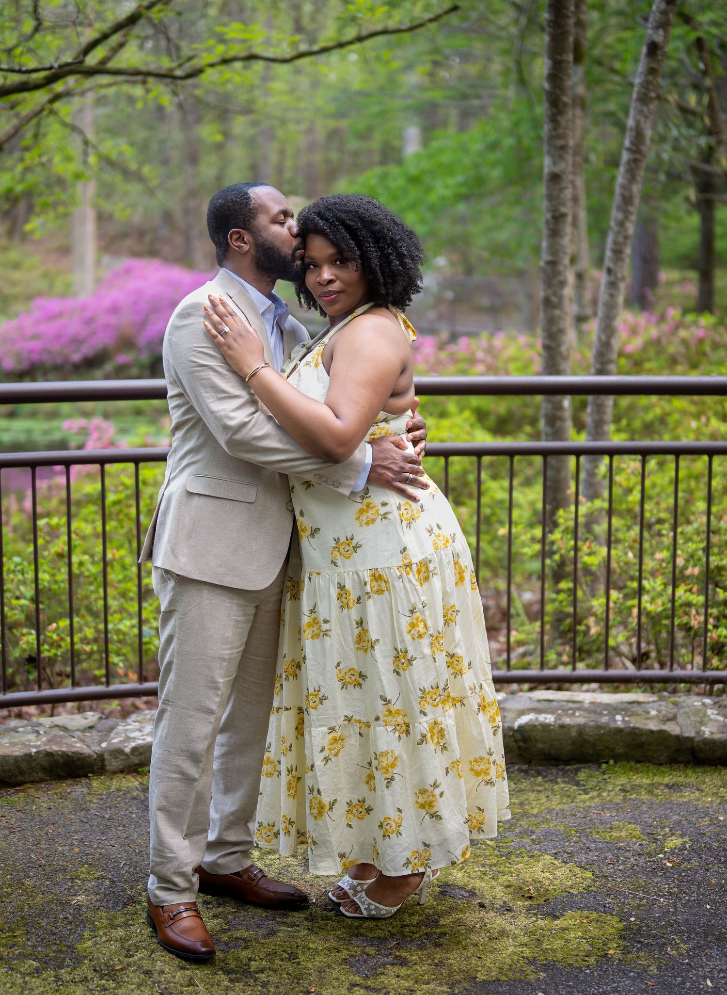 Couple embracing tenderly on a mossy terrace surrounded by blooming azaleas in a lush garden.