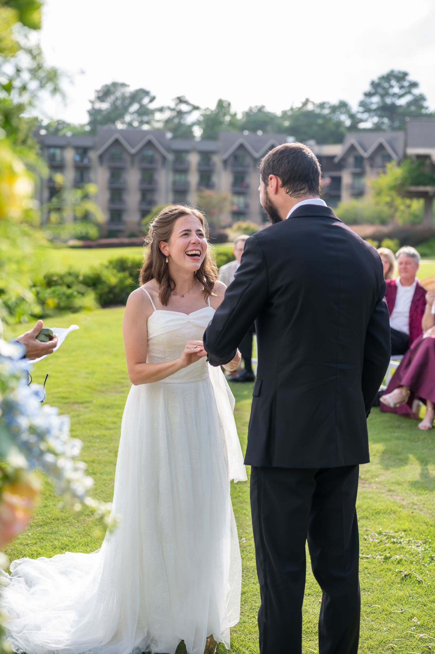 A joyful outdoor wedding ceremony takes place on a lush lawn with apartment buildings visible in the background.