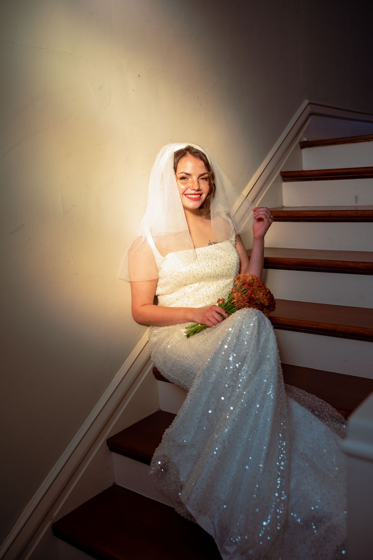 A person wearing a sparkling white wedding gown sits dramatically on a staircase in a shadowy setting.