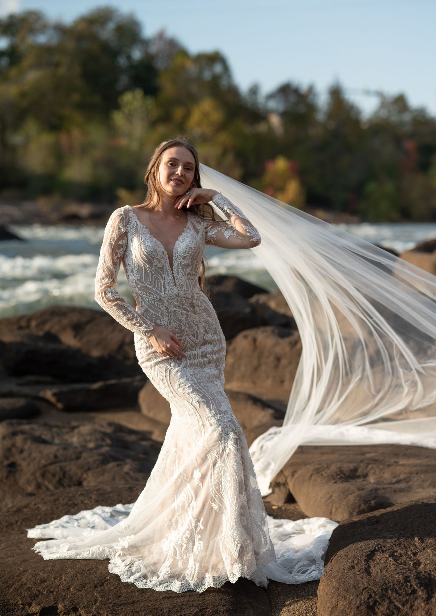 Bride in long-sleeve lace gown poses gracefully on rocky terrain with veil flowing in the breeze.